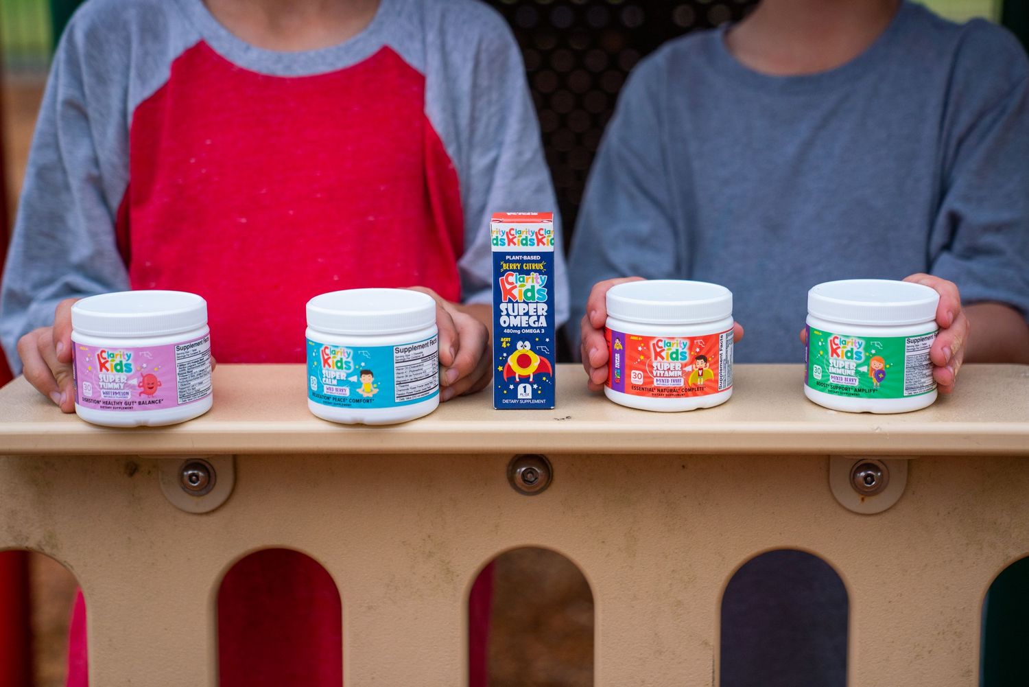 Two children holding colorful kids supplement containers on a wooden shelf outdoors.