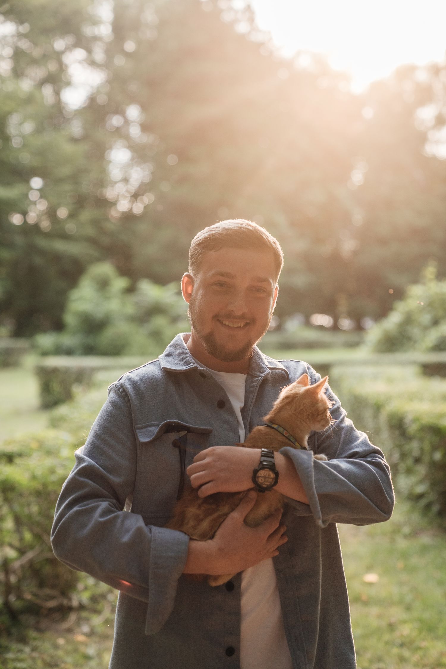 Smiling man holding an orange tabby cat outdoors in a sunny green park wearing a gray jacket.