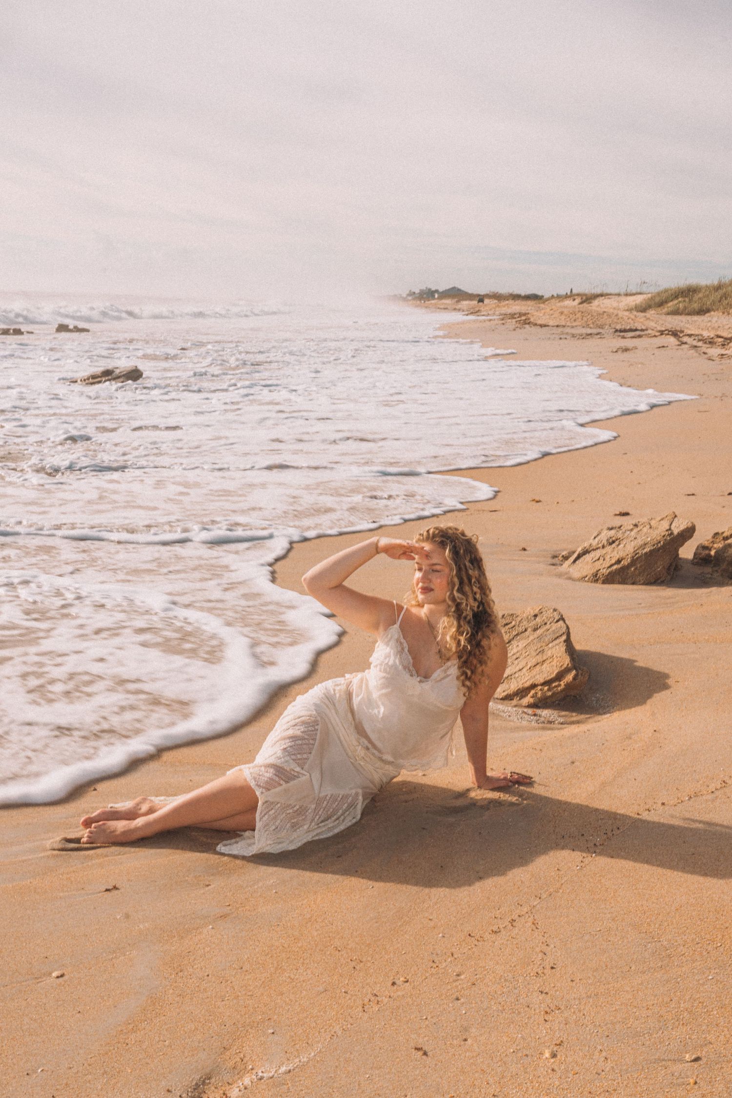 Woman in white lace dress sitting on sandy beach with ocean waves nearby.