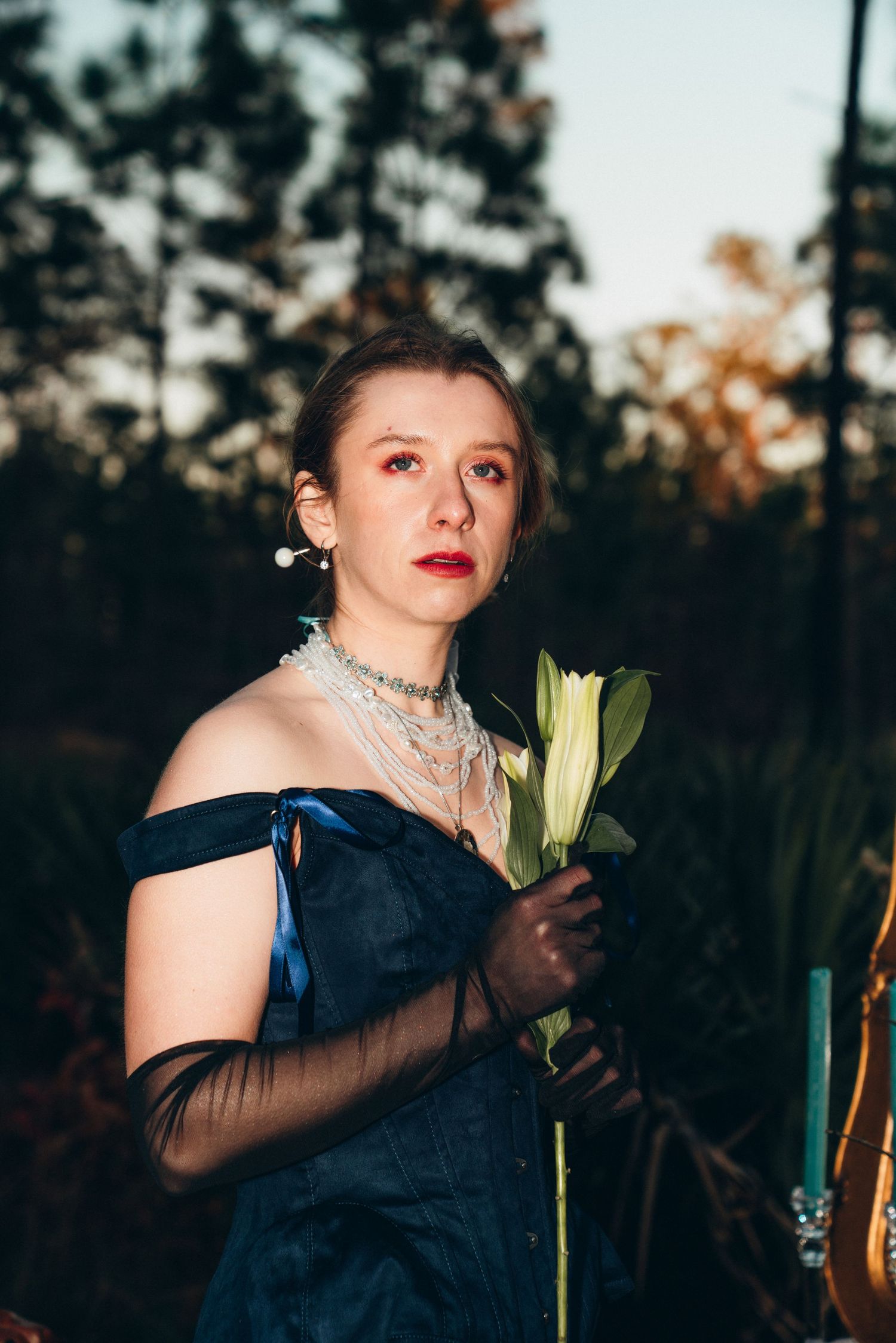 Woman in blue off-shoulder gown holding white lily flower, wearing pearl necklace, posing outdoors at dusk.