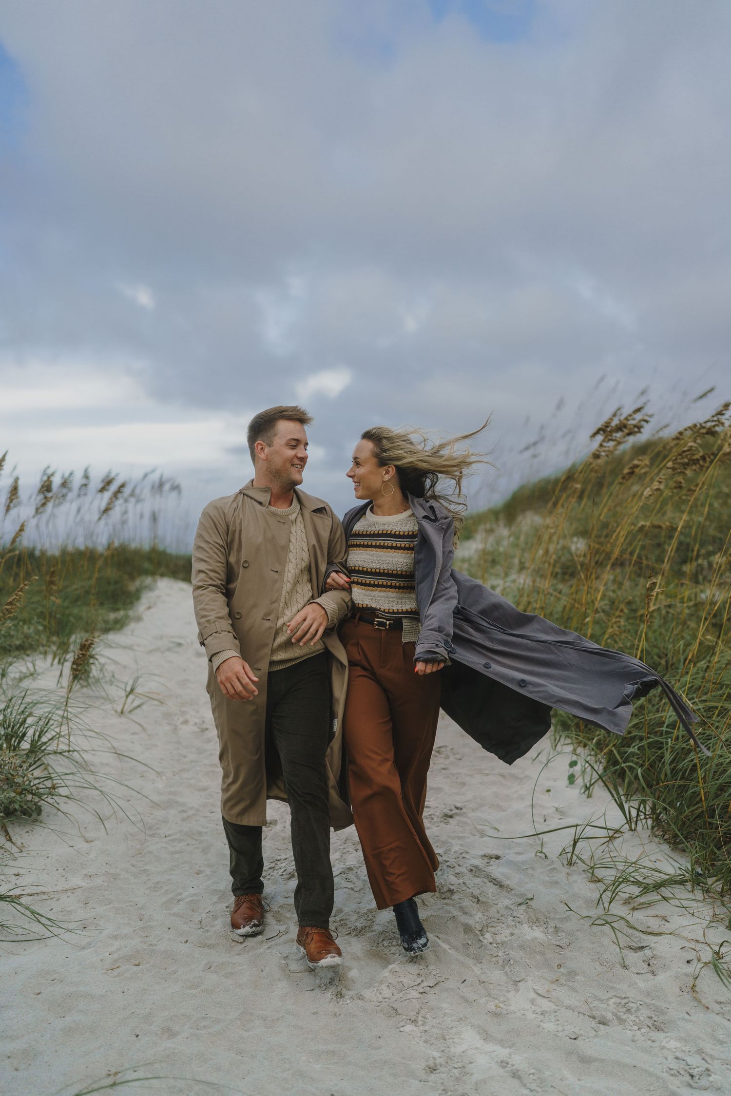 Couple walking and embracing on a windy beach with sea grass and sand dunes in the background.