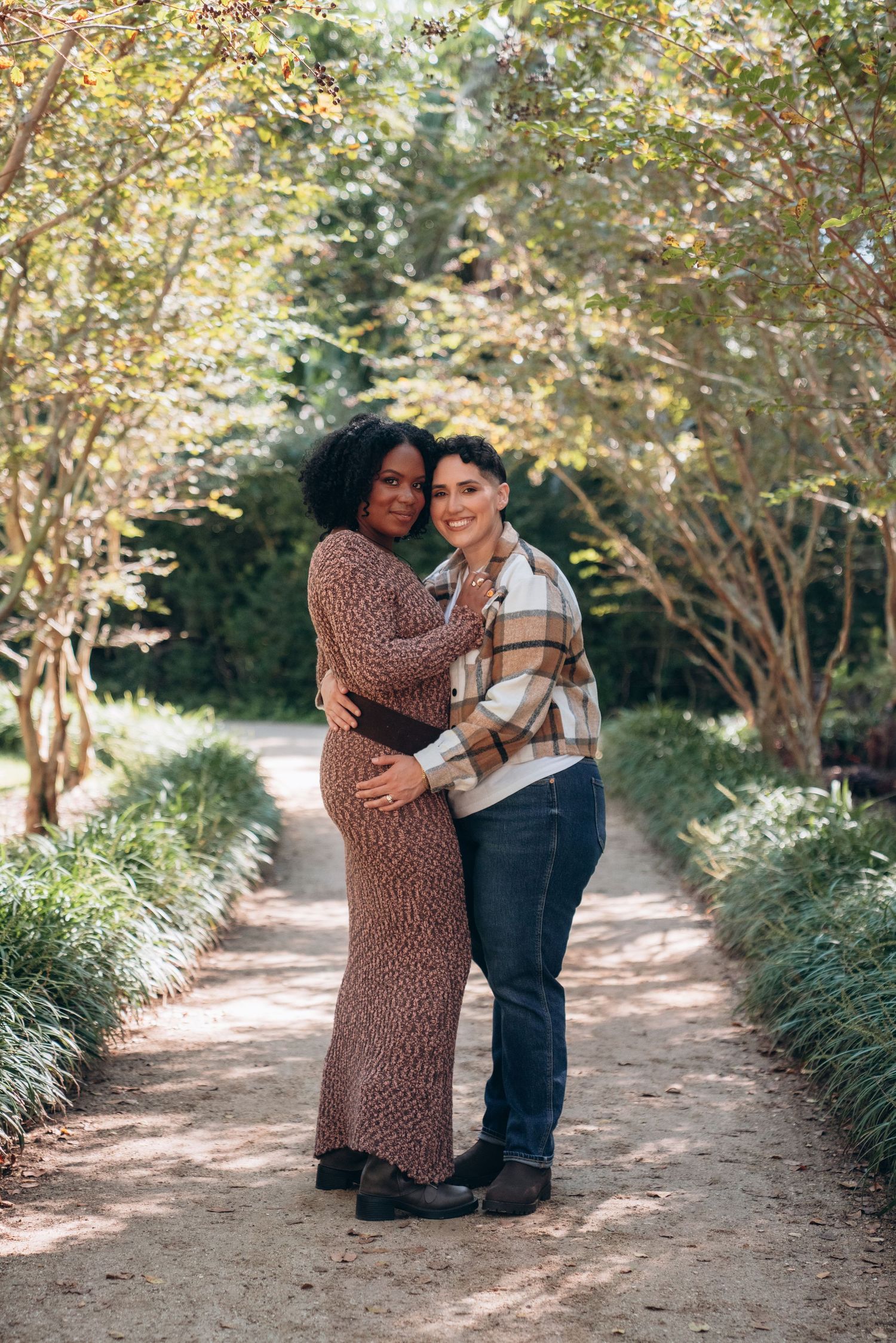 Two people embrace while standing on a garden path surrounded by autumn foliage and trees.