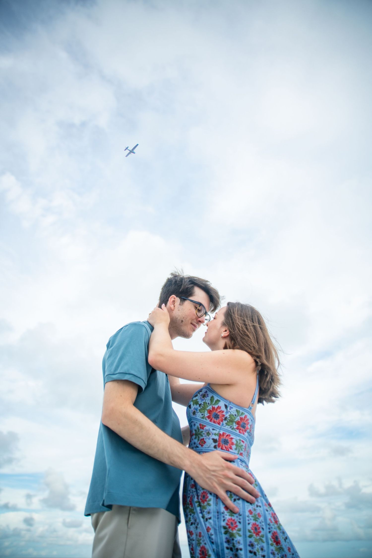 A romantic couple embraces on the beach with a small airplane visible in the blue sky above.
