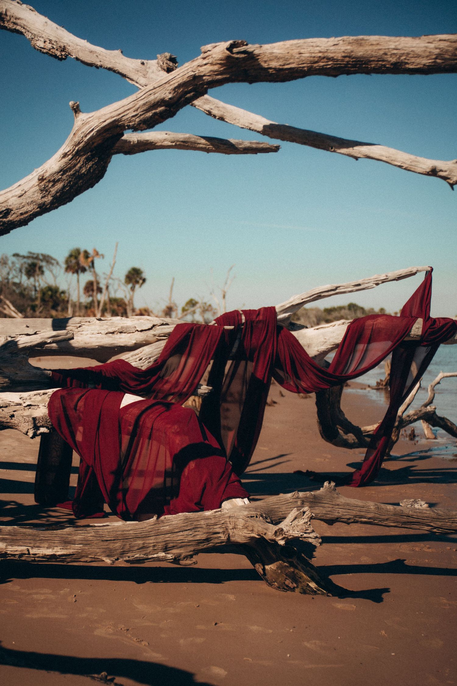 Red fabric draped over driftwood on a beach against a bright blue sky.