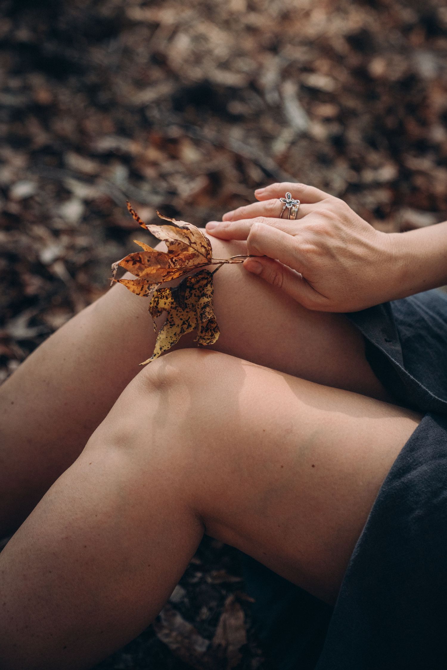 A delicate hand rests on bare knees holding autumn leaves against a dark forest floor background.