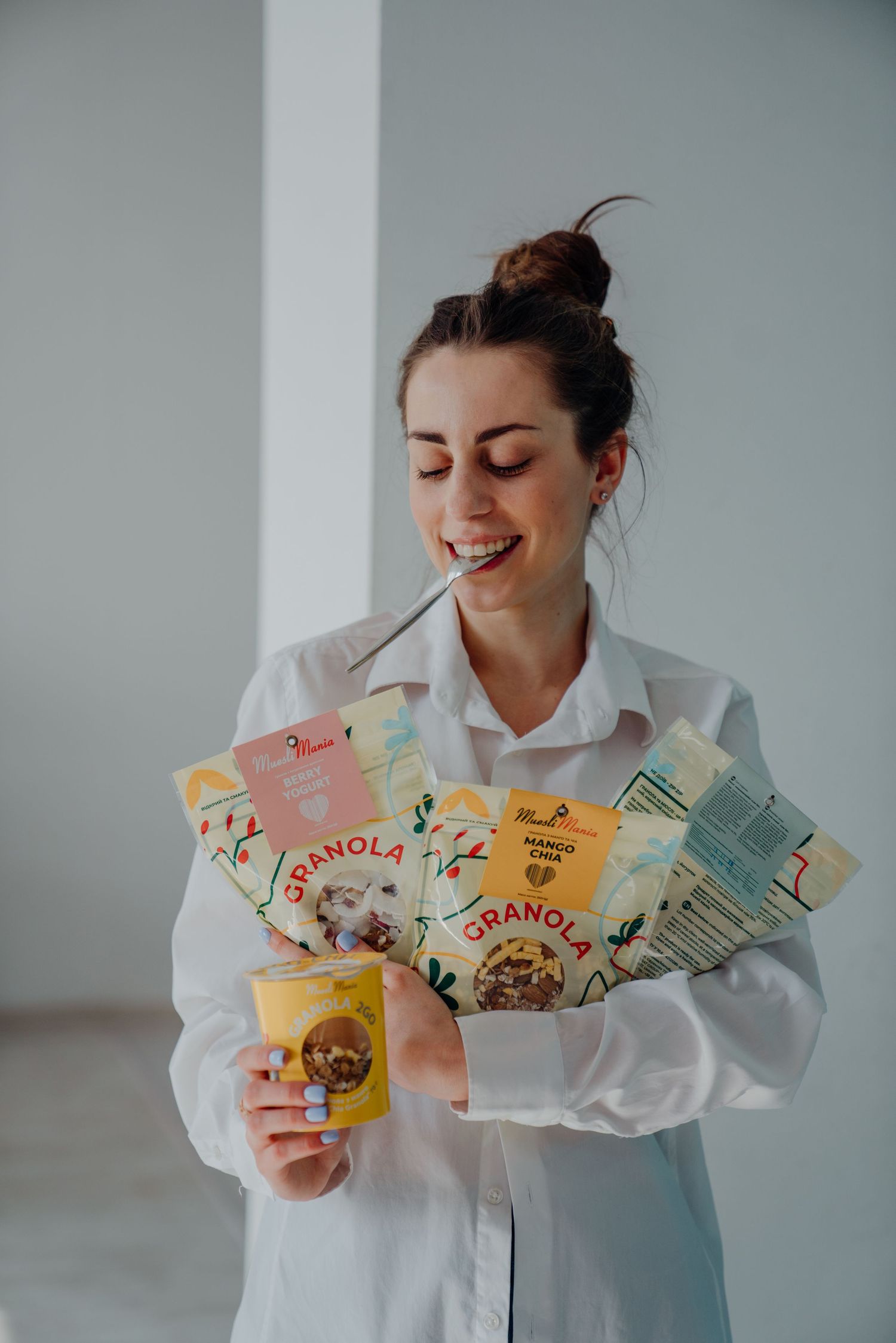Happy woman in white shirt holding granola packages and a yellow smoothie cup indoors.