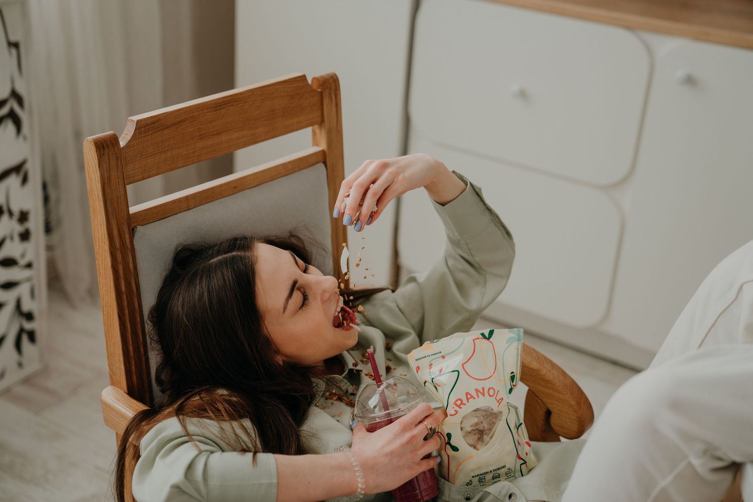 Young woman relaxing on a wooden chair, playfully eating a snack while holding a decorative bag indoors.
