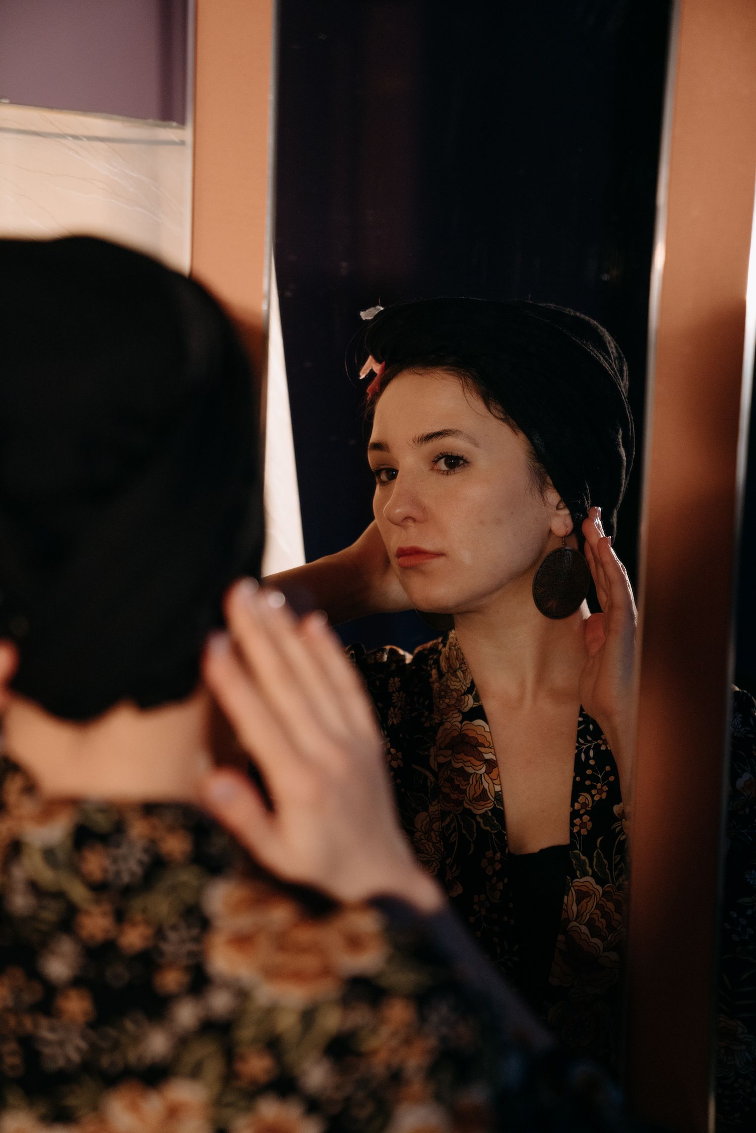 Woman in black turban and floral dress adjusting earring while looking at her reflection in a mirror.