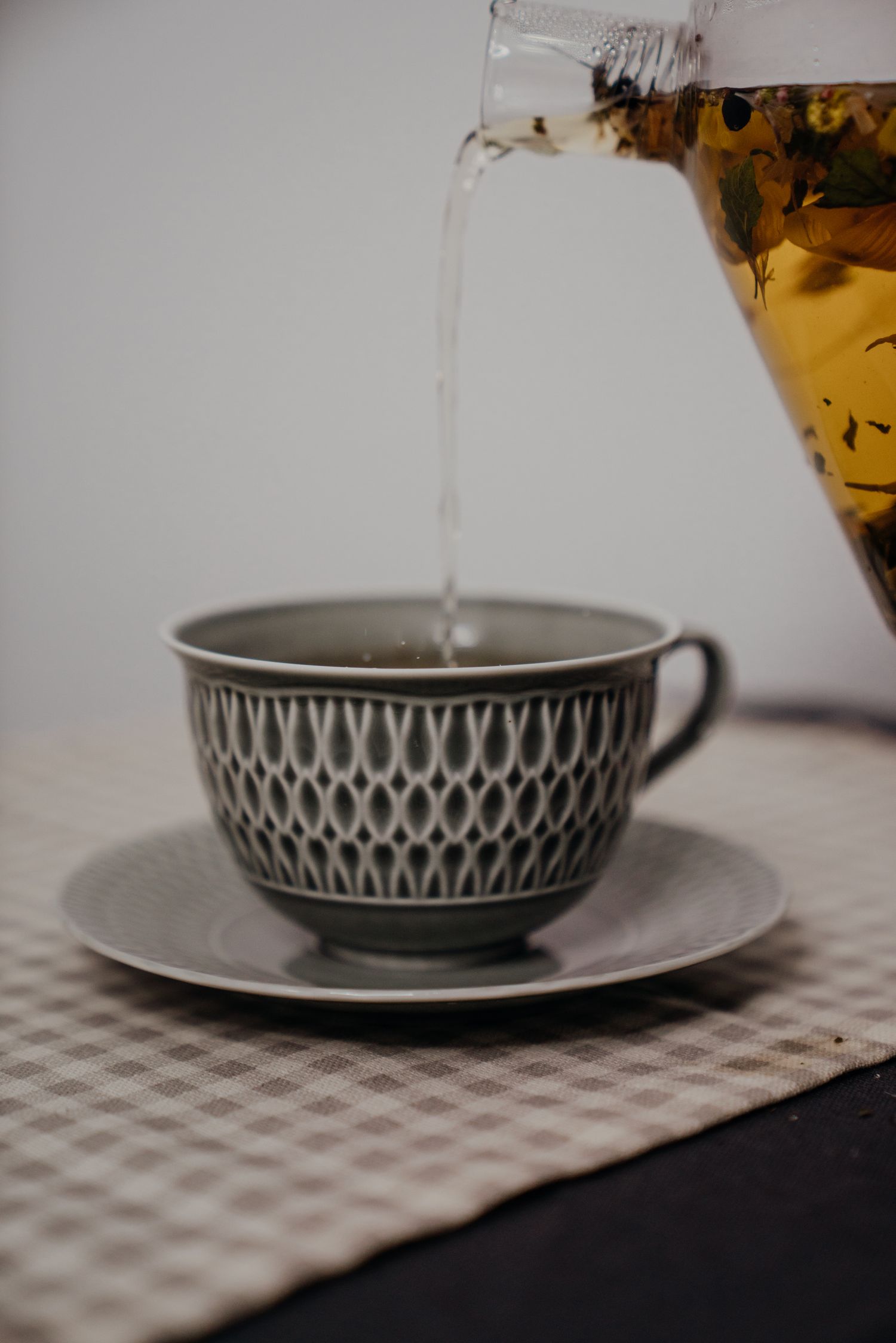 Herbal tea being poured from a glass teapot into a decorative gray ceramic cup on a saucer.
