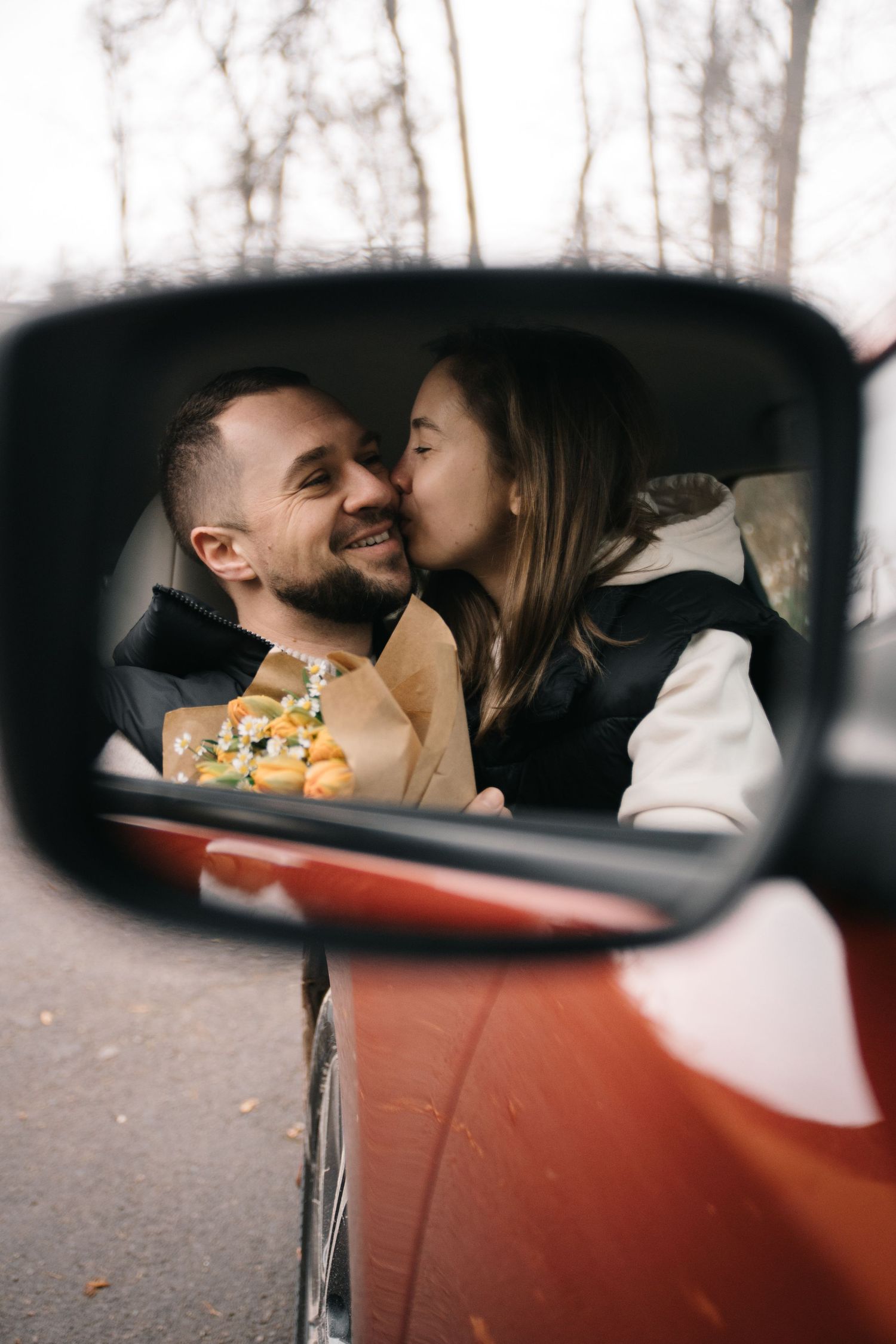 Couple kissing inside car reflected in side mirror, woman holding small yellow bouquet.