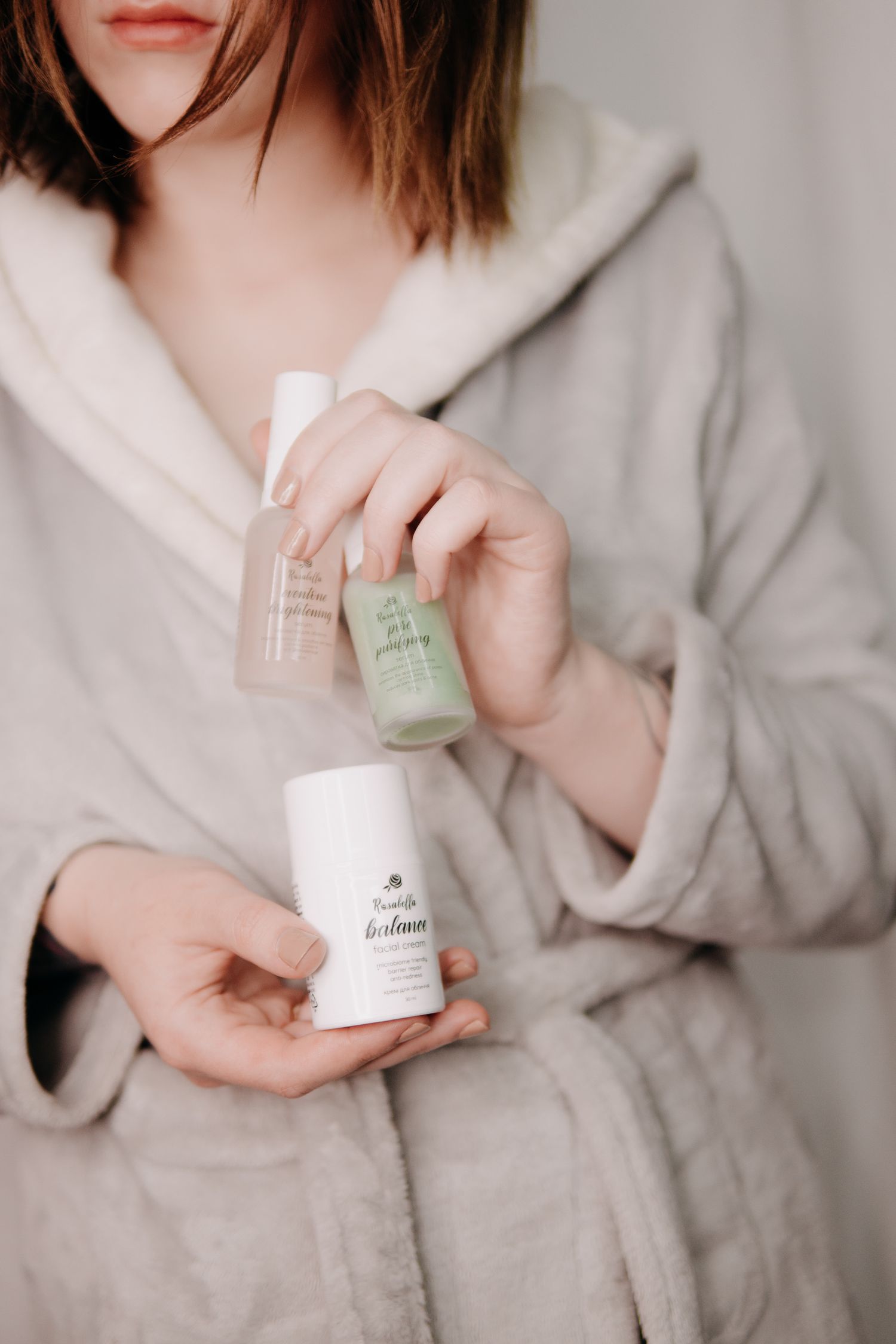 Woman in cozy gray robe holding three skincare products including serums and a deodorant in soft natural light.