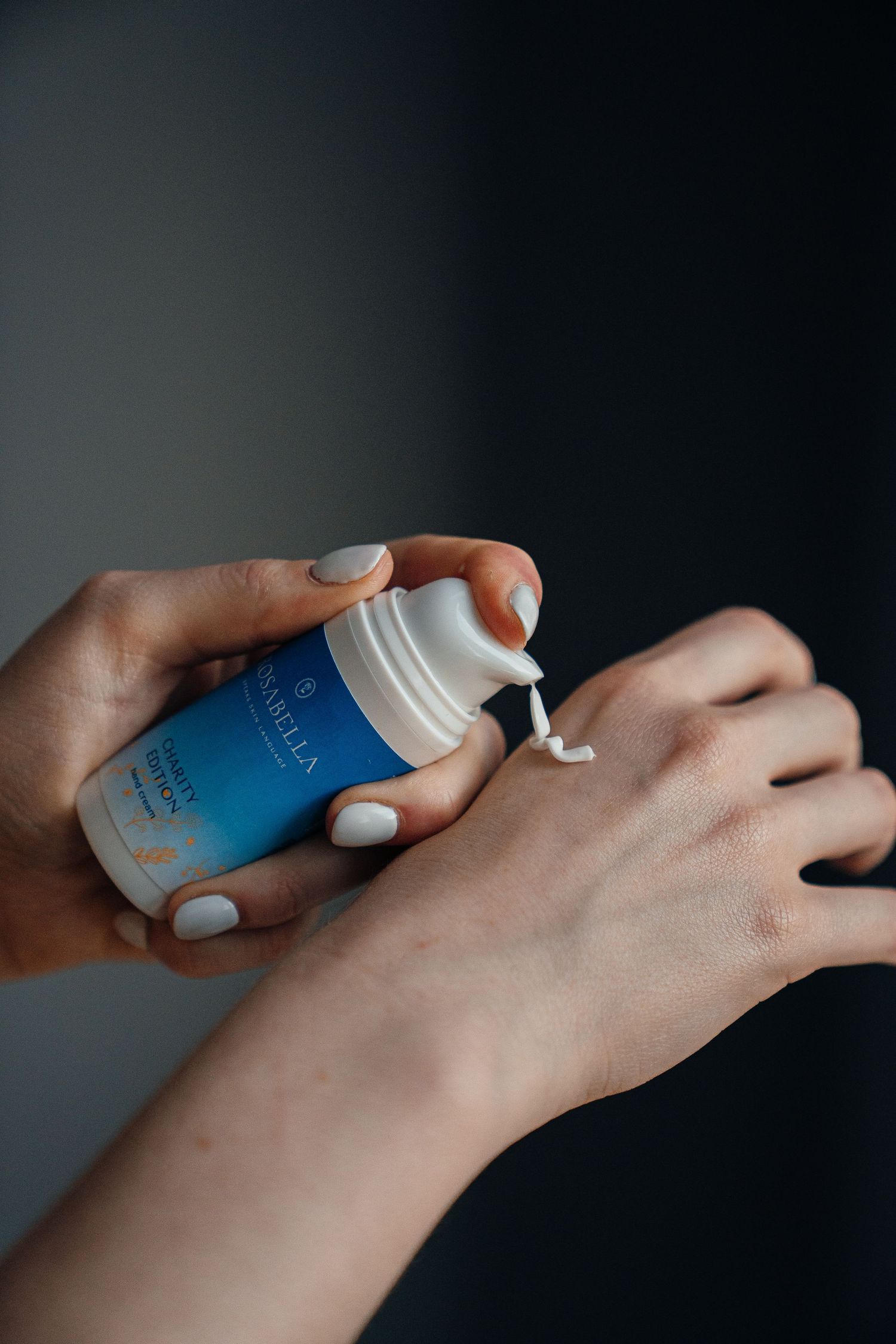 Hands dispensing white cream from a blue Varblla skincare bottle onto skin against dark background.