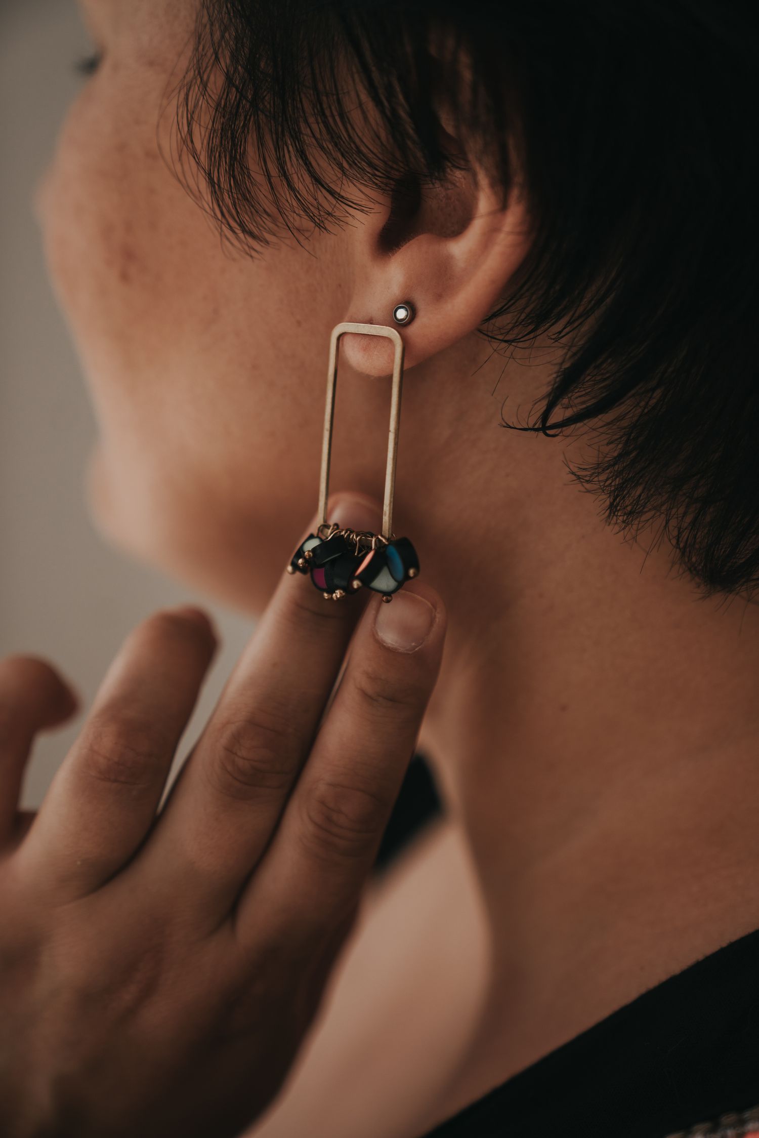 Woman wearing a modern rectangular gold earring with dark gemstones, held up to her ear.
