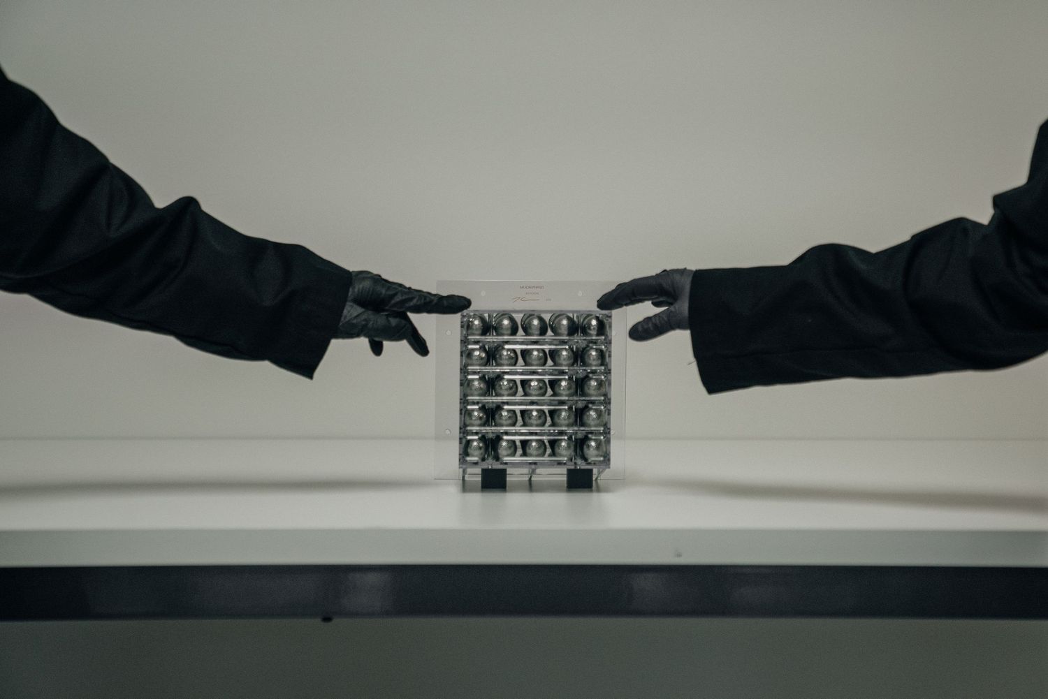 Two gloved hands pointing at a metallic battery cell module on a white table in a dramatic studio setting.