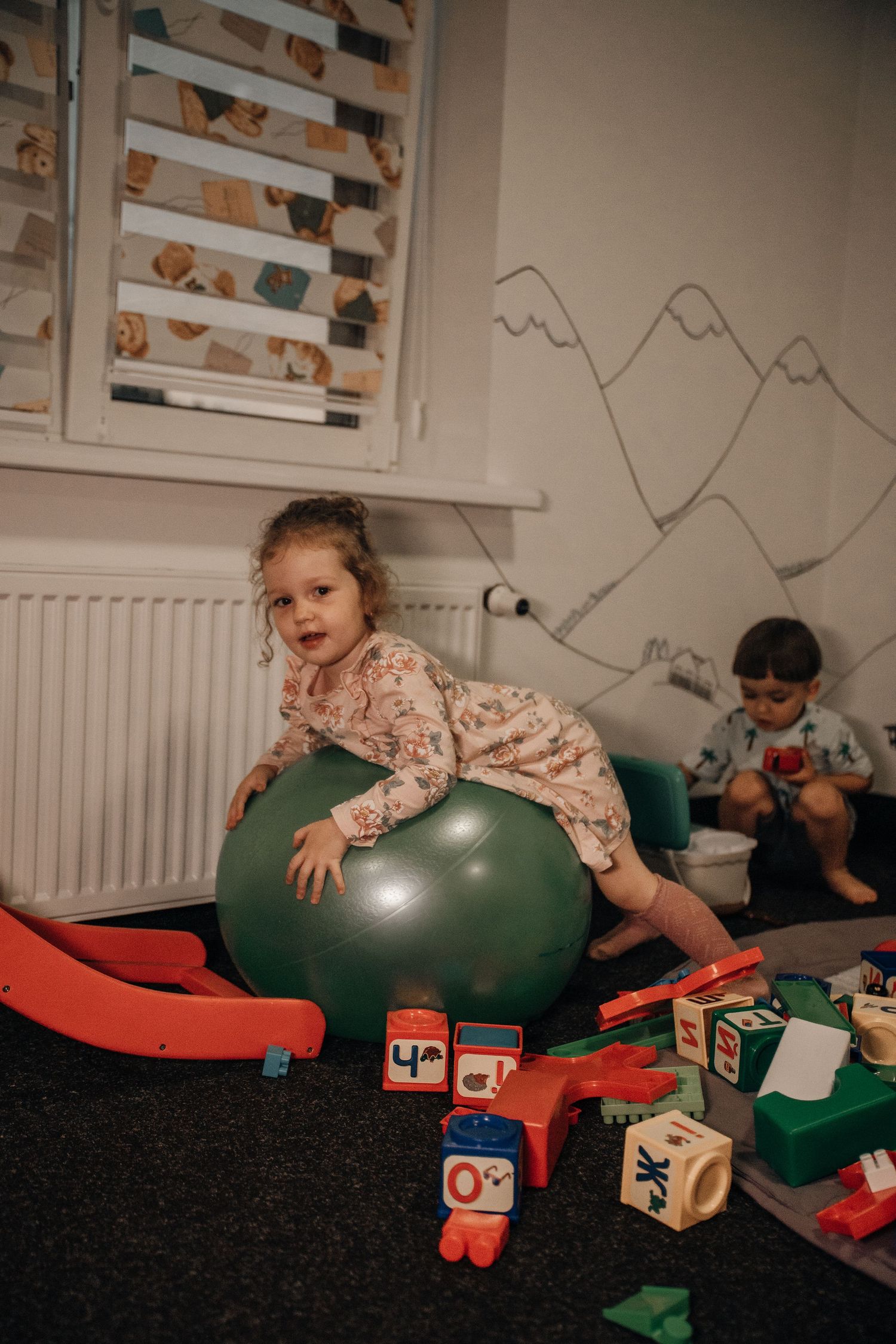 Young toddler girl lying on a green exercise ball surrounded by colorful wooden toys in a playroom.