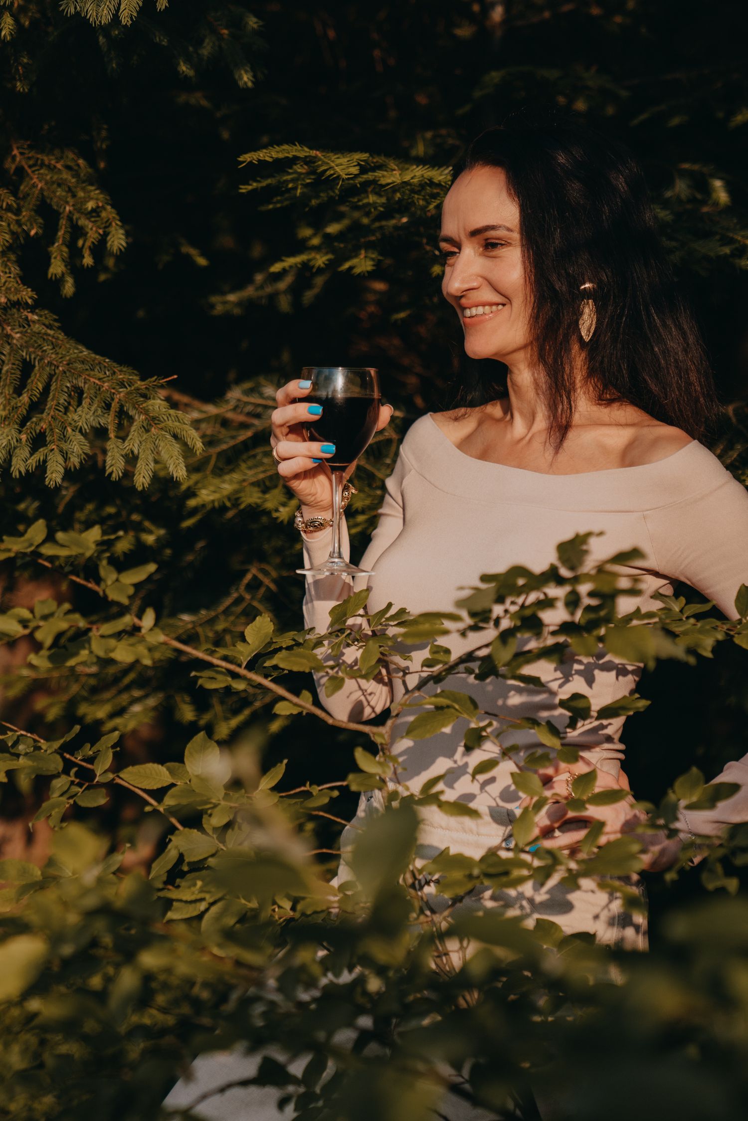 A smiling woman holding a glass of red wine outdoors among lush green trees in warm sunlight.
