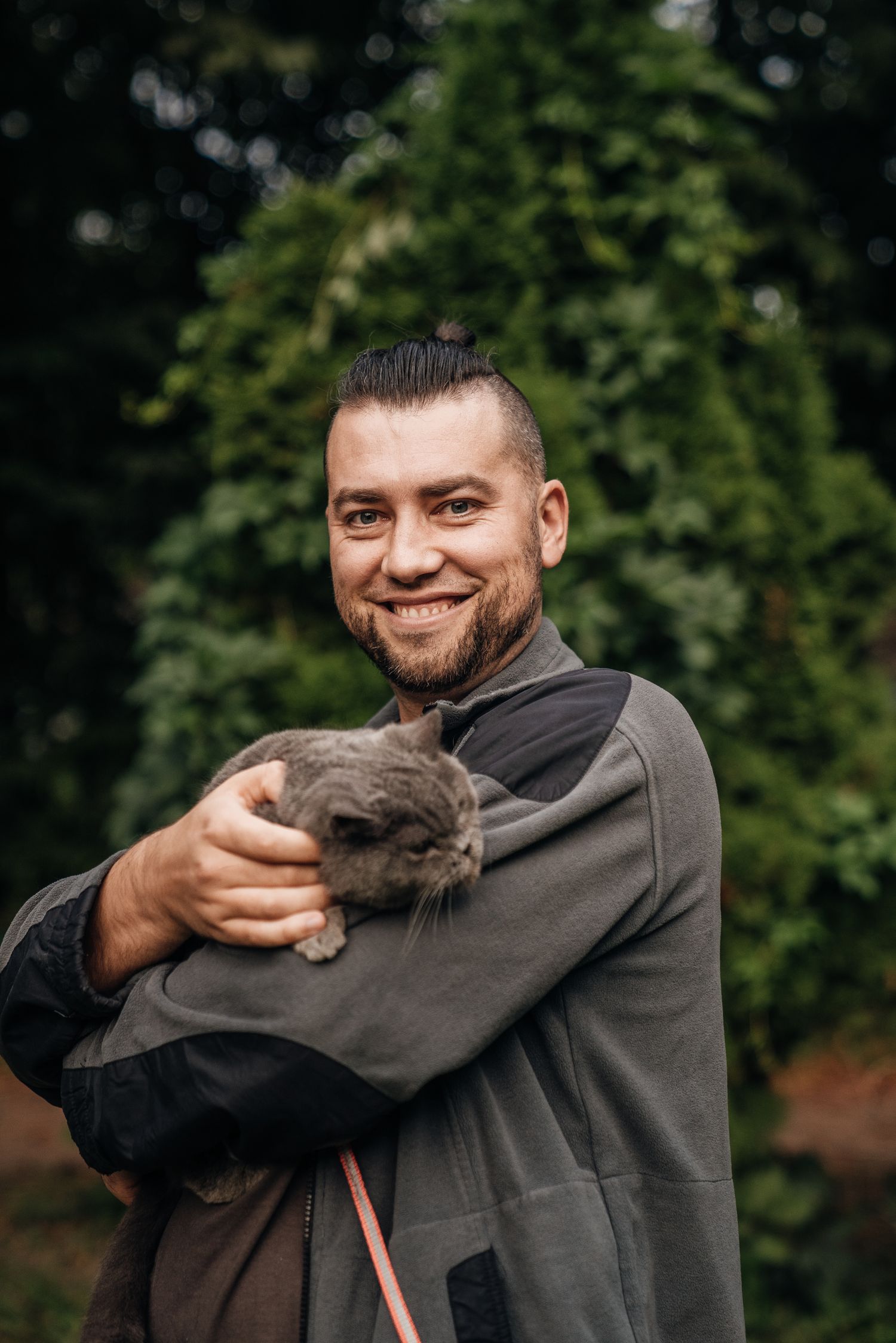 Smiling bearded man holding a small gray rabbit outdoors with green trees in background.