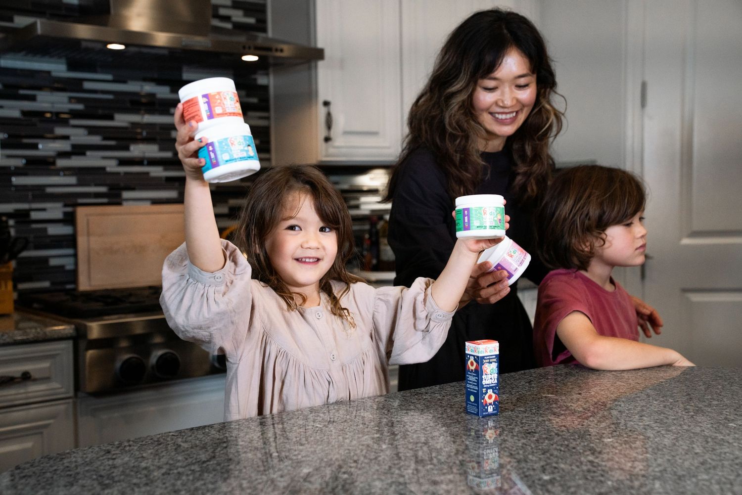 A mom and two young children happily hold up colorful yogurt cups in a modern kitchen.
