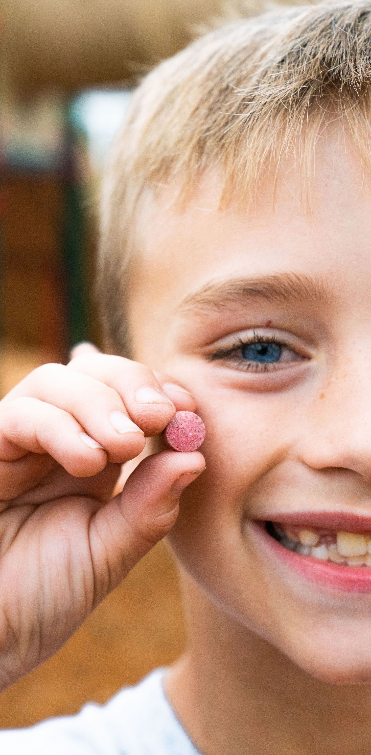 Smiling blonde boy with blue eyes holding up a small pink candy close to his face outdoors.
