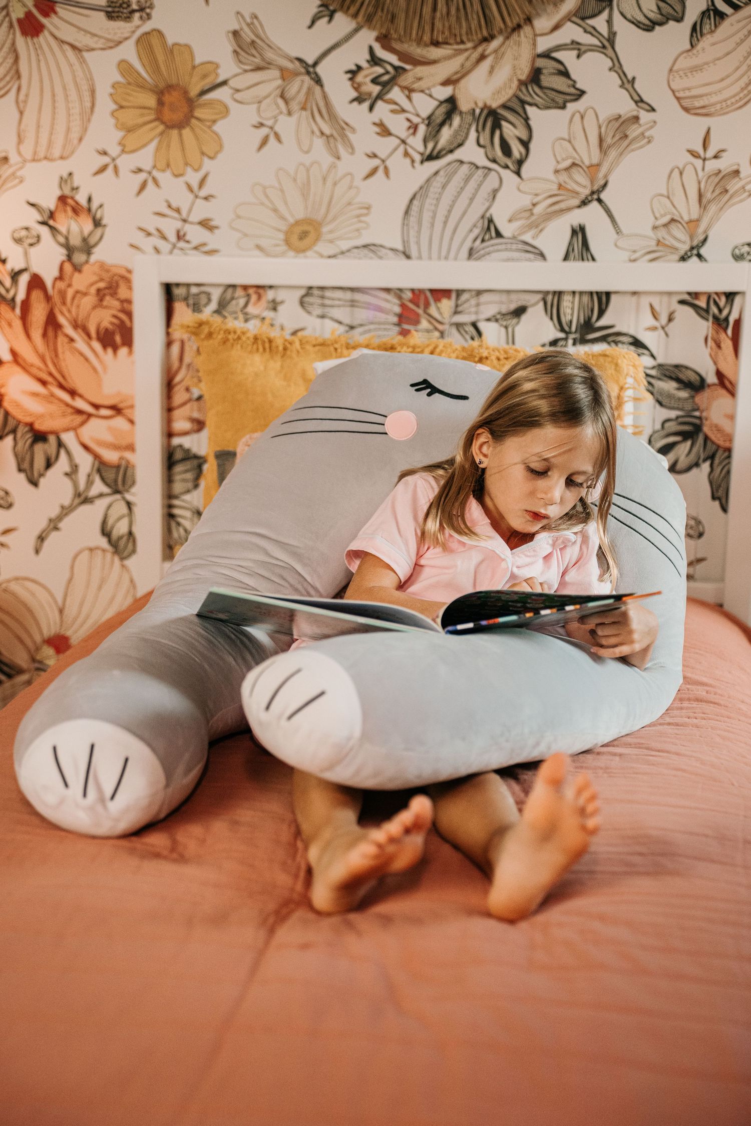Young girl reading a book while leaning against a large grey cat stuffed animal pillow on bed.