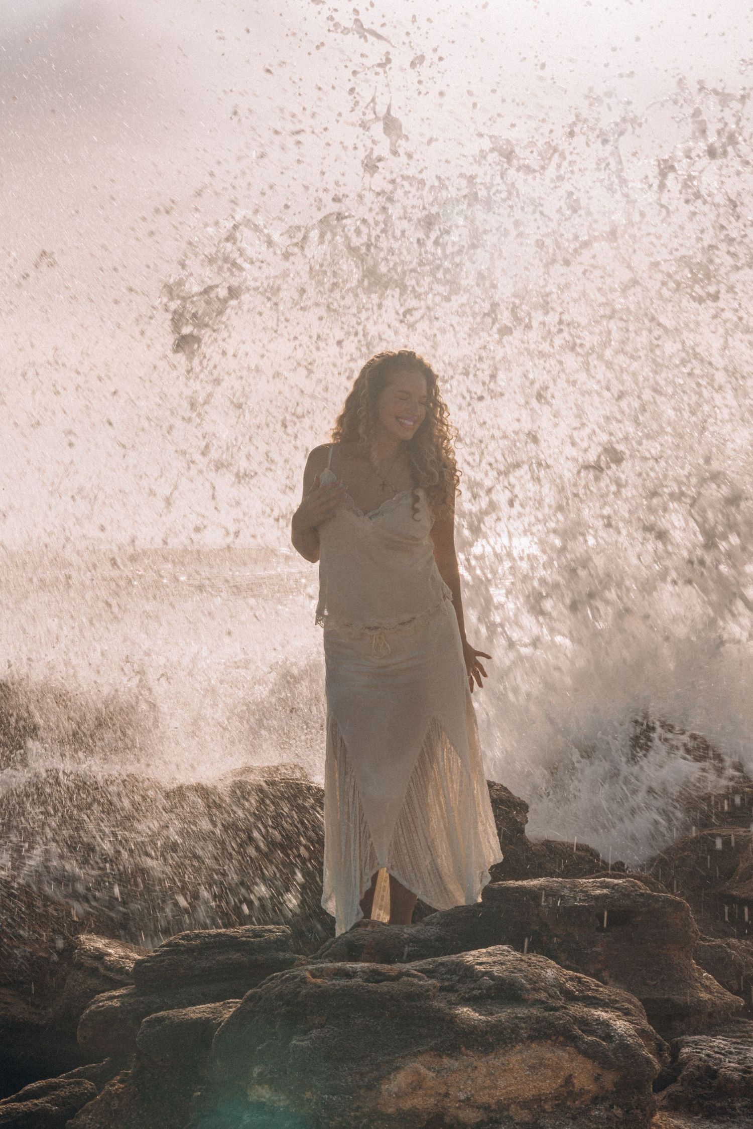 Woman in white dress standing on rocks with ocean waves crashing dramatically behind her in golden light.