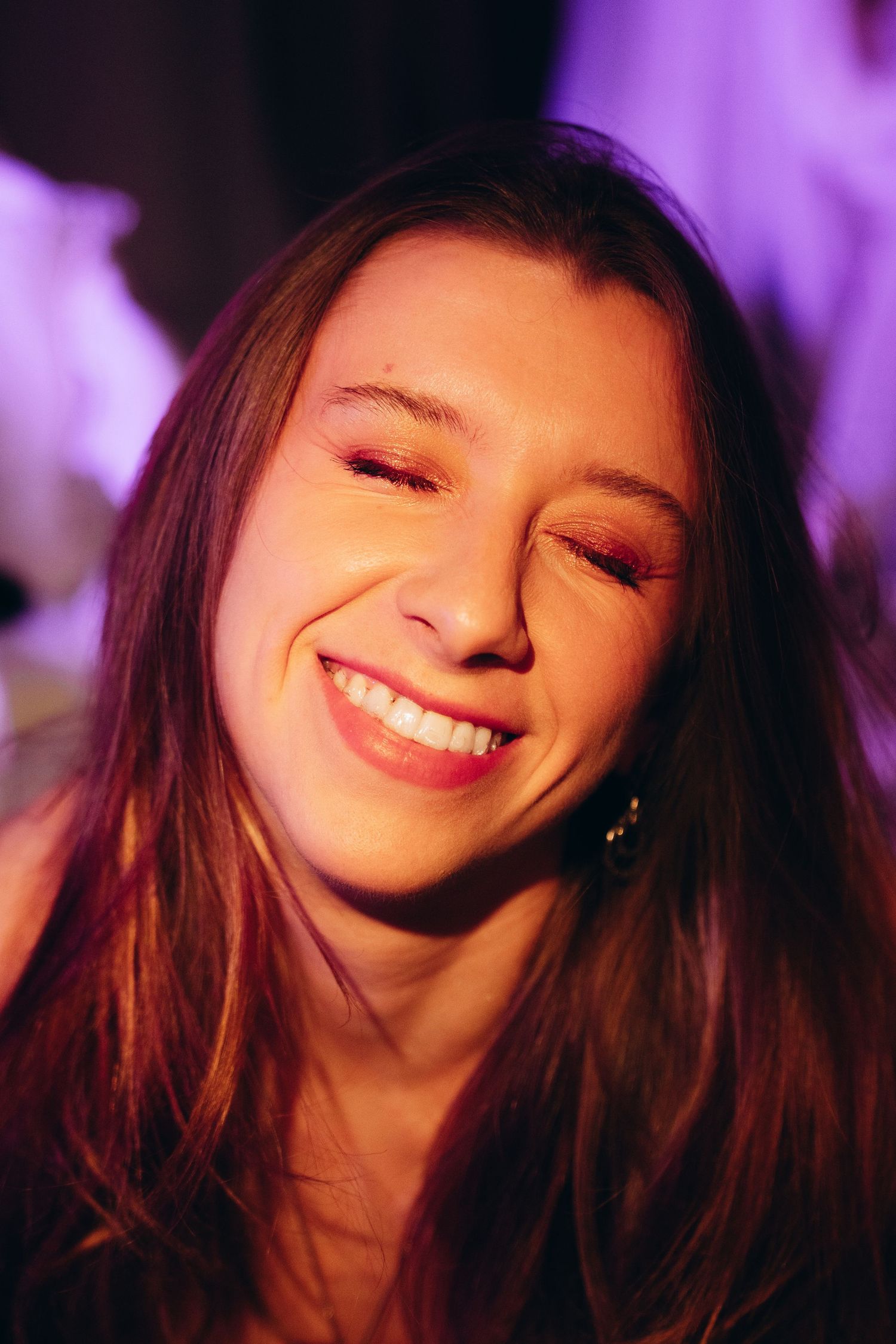 Young woman smiling with eyes closed under purple party lights, wearing earrings and natural makeup.