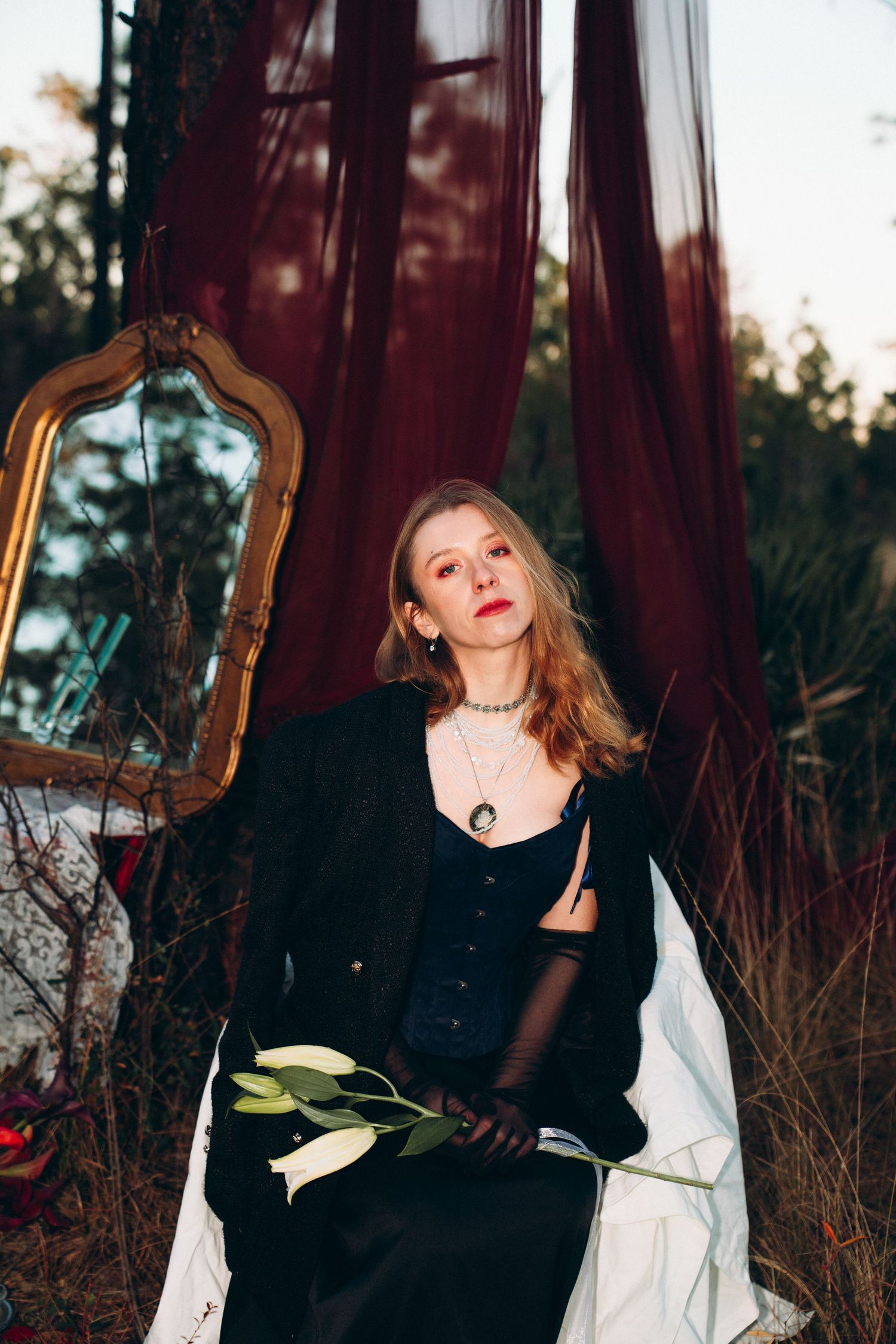 Blonde woman in black jacket and dress holds white flower beside gold mirror with burgundy drapes outdoors.