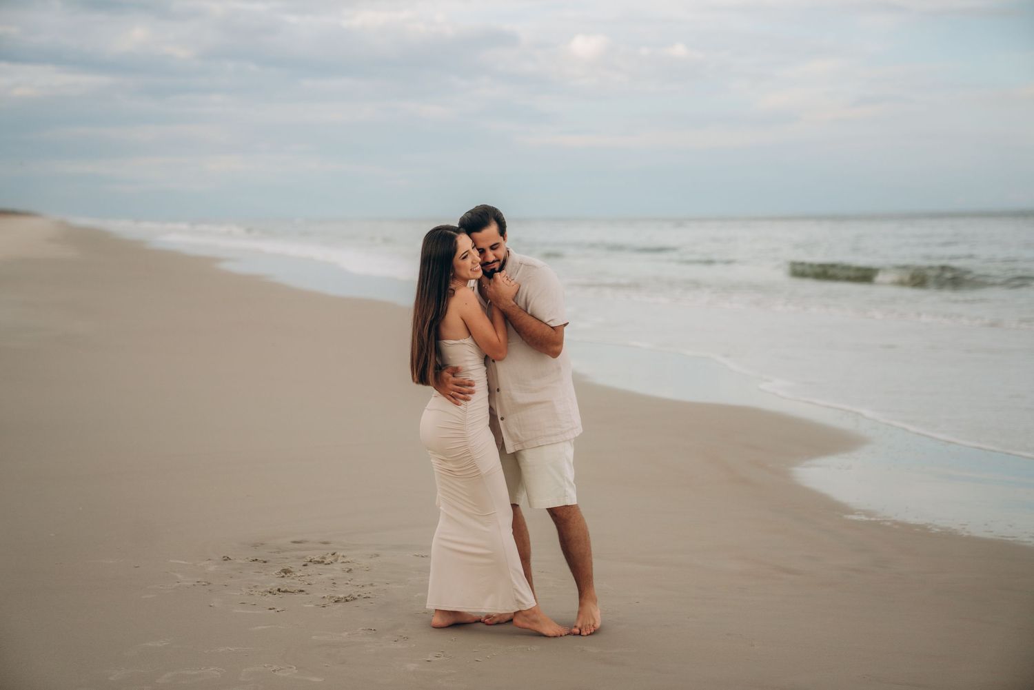 Couple walking together on beach with waves and cloudy sky in background.