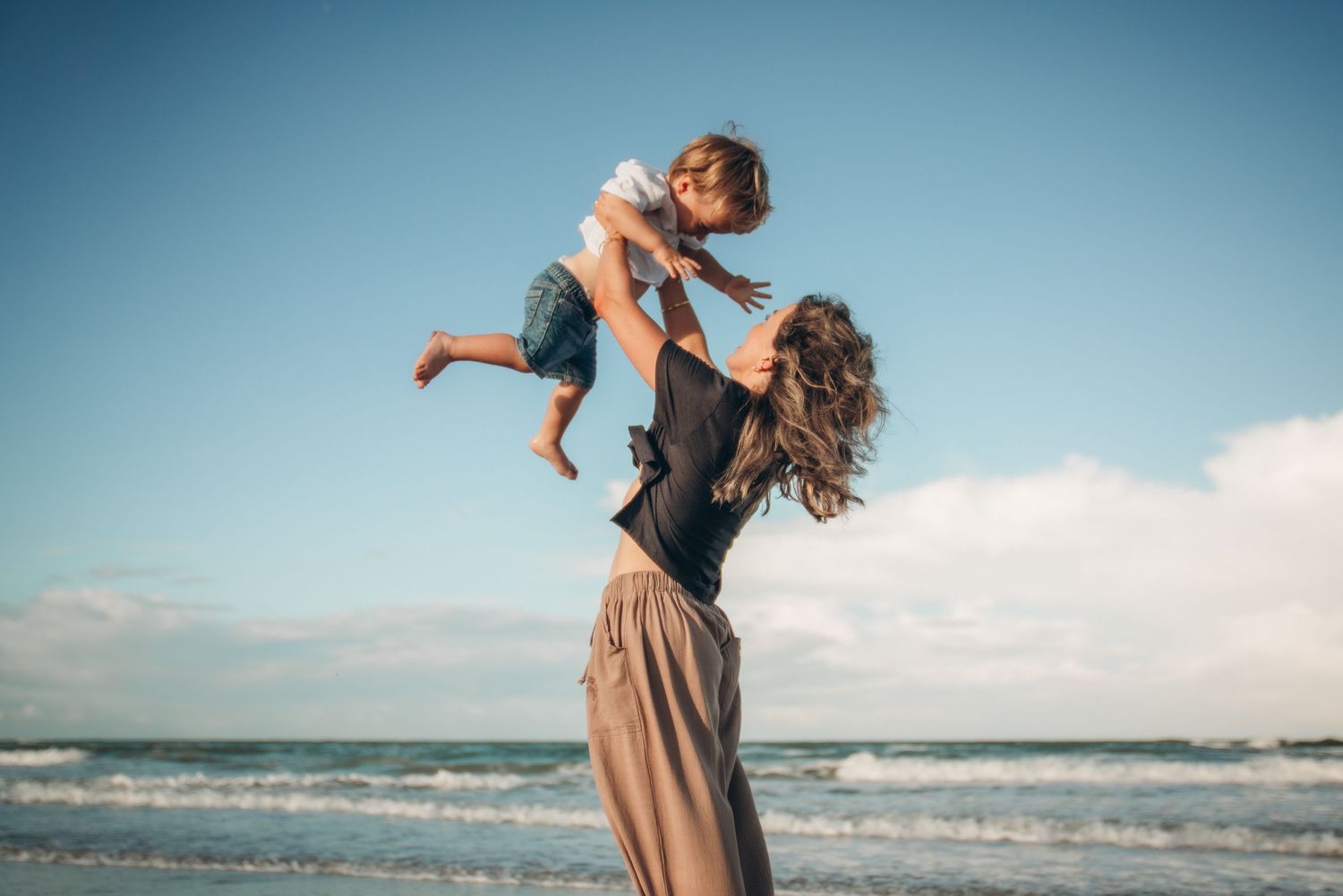 A joyful mother lifting her happy toddler high in the air on a sunny beach with blue skies and ocean waves.