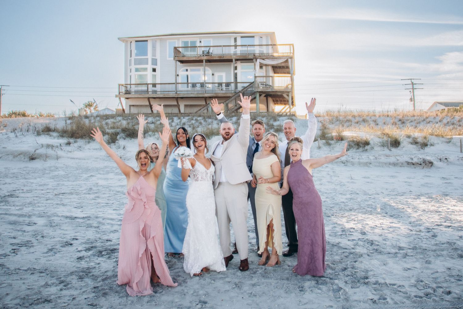 Wedding party celebrating on beach with raised arms, bride in white gown, bridesmaids in colorful dresses, beach house backdrop.