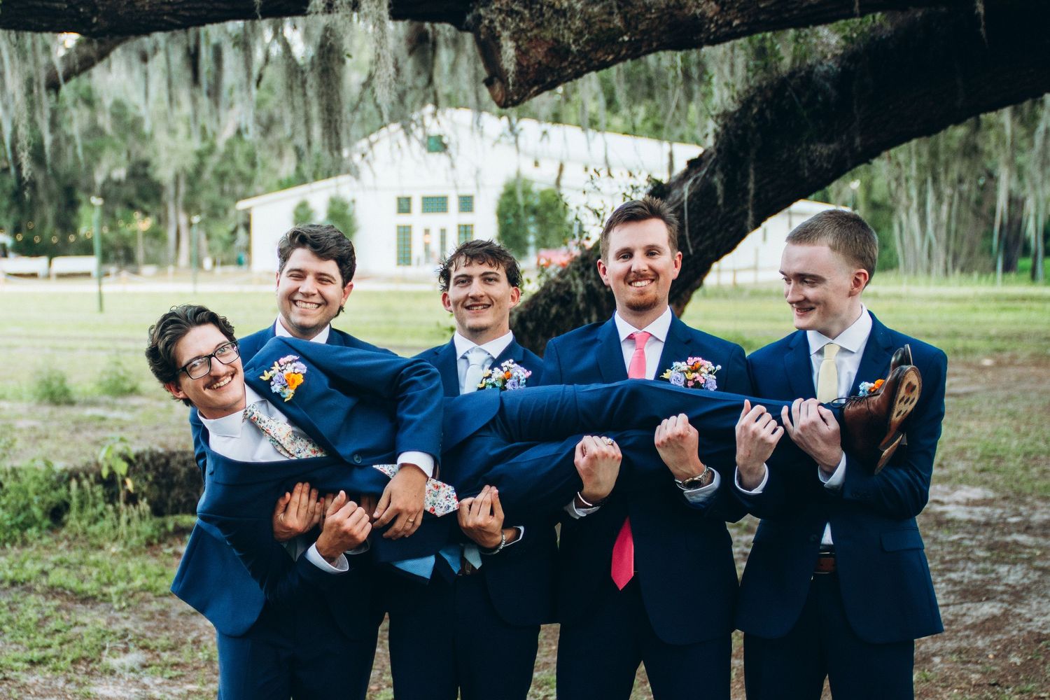 Five groomsmen in navy blue suits pose playfully under a large oak tree at an outdoor wedding venue.