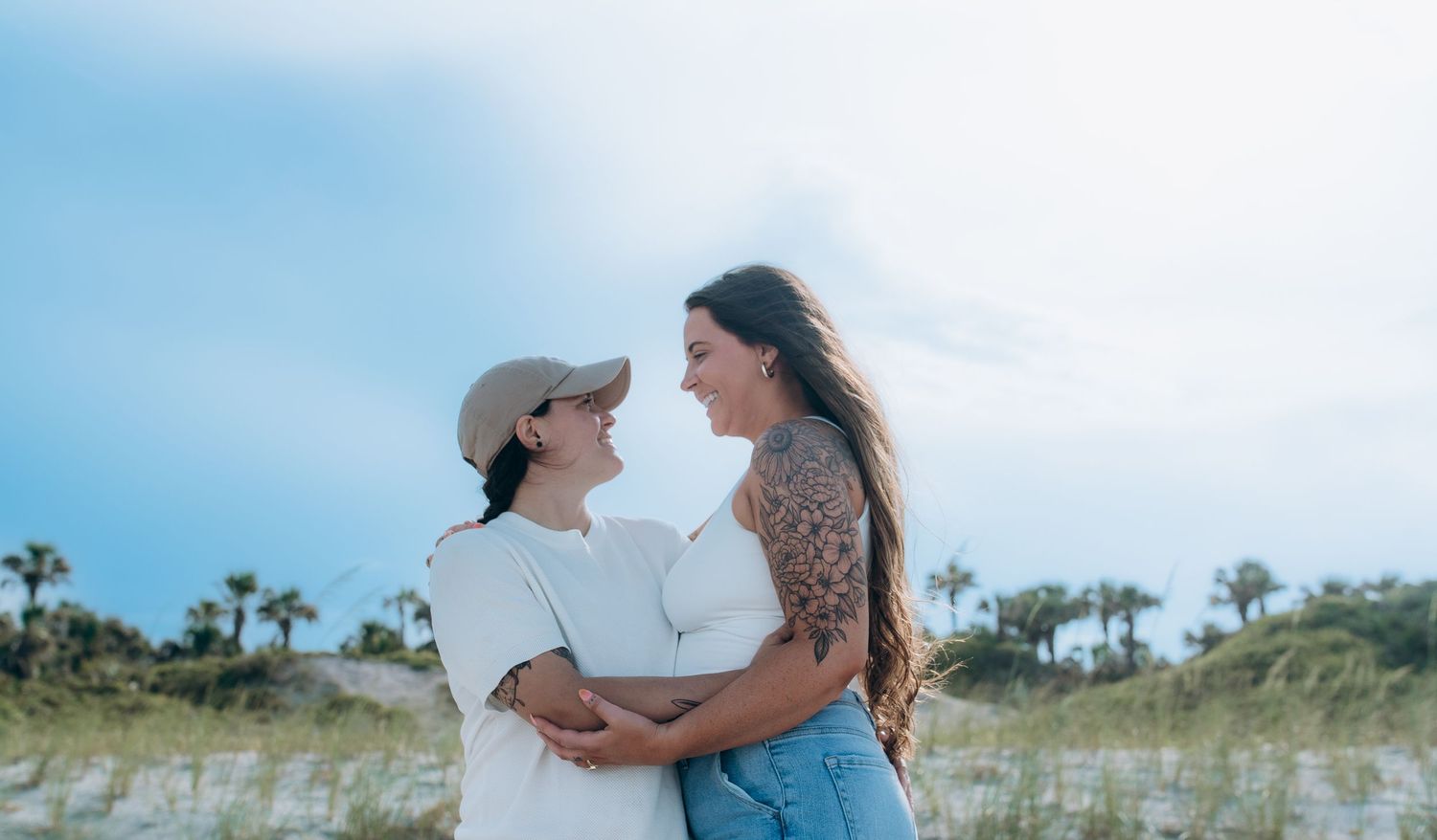 Couple embracing on beach with palm trees and blue sky in background.