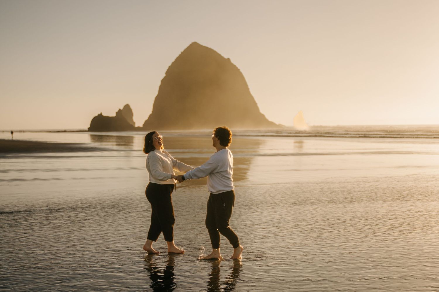 Two people standing in shallow water at sunset with coastal rocks in the background.