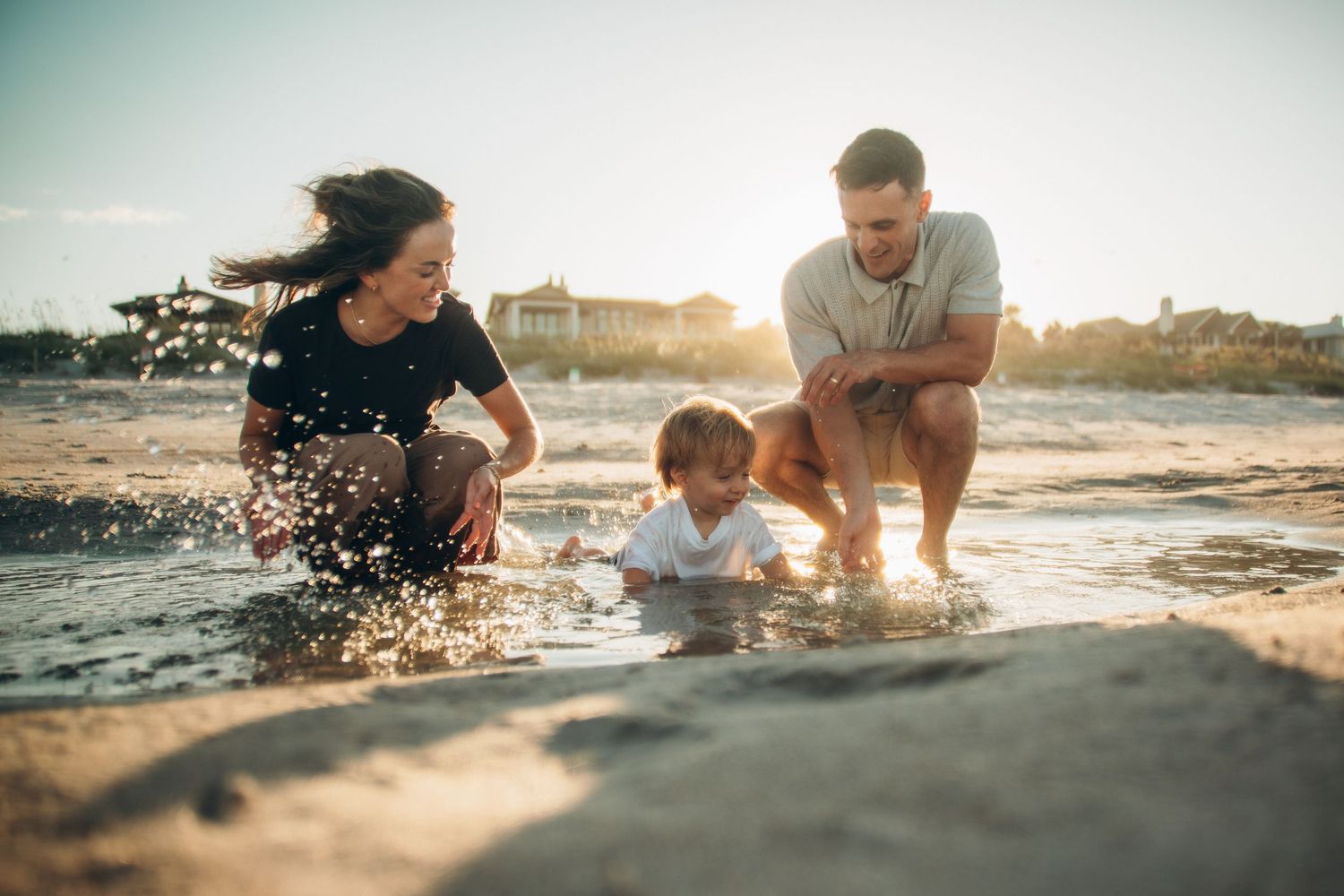 Family playing at beach shoreline with toddler splashing in waves during golden sunset hour.