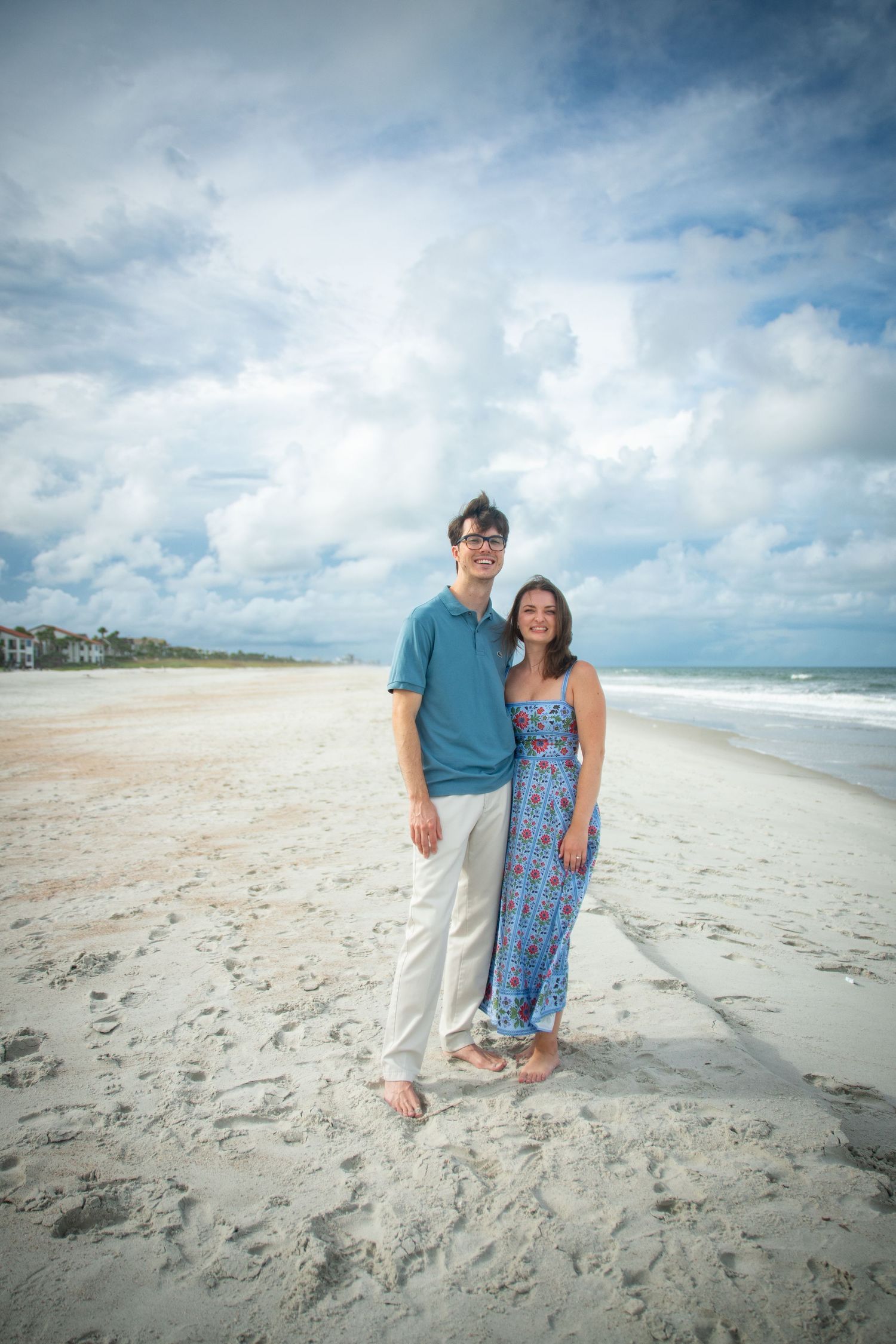 A couple stands together on a sunny beach with white sand and blue skies in the background.