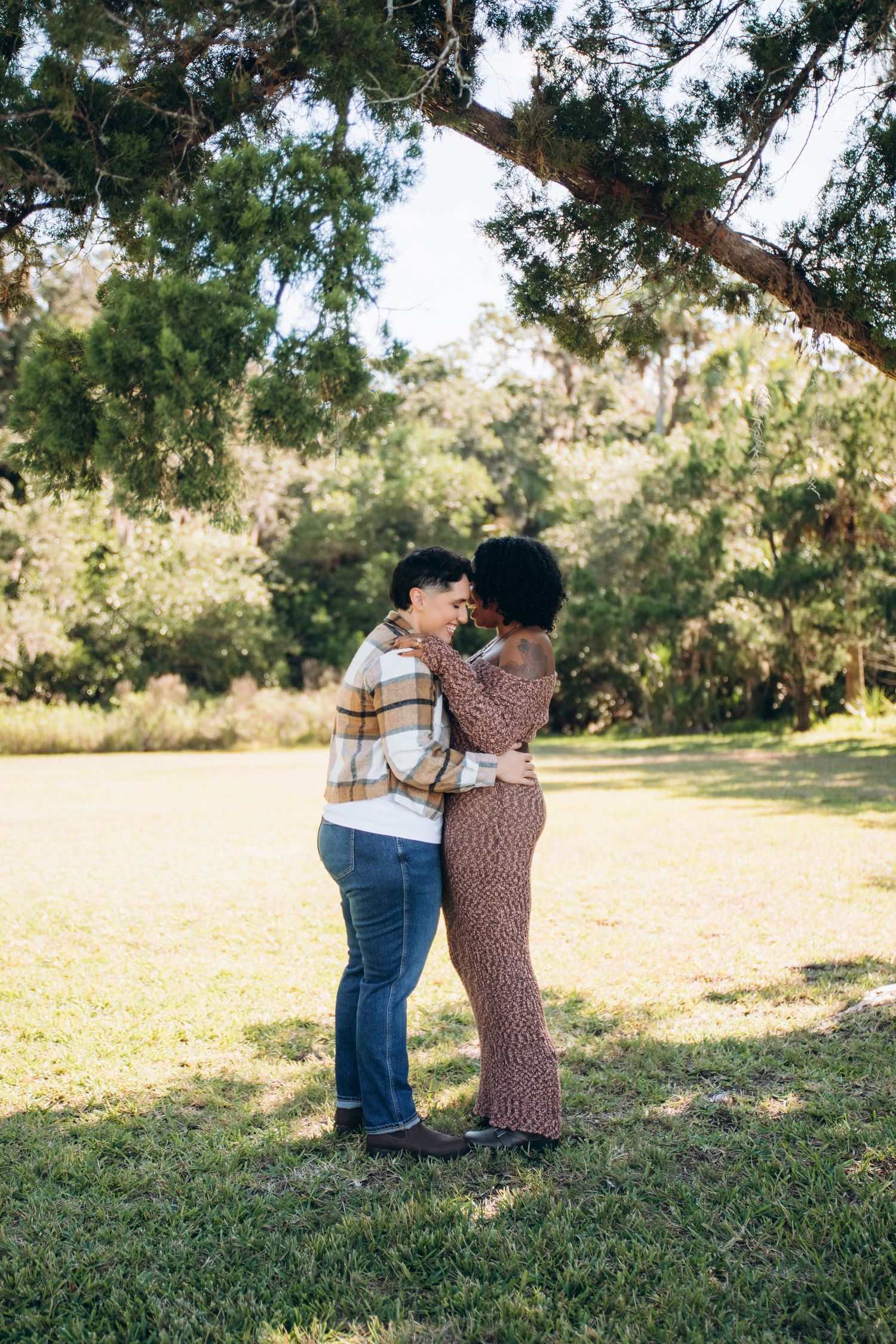 A couple embraces while walking through a sunlit park with trees in the background.
