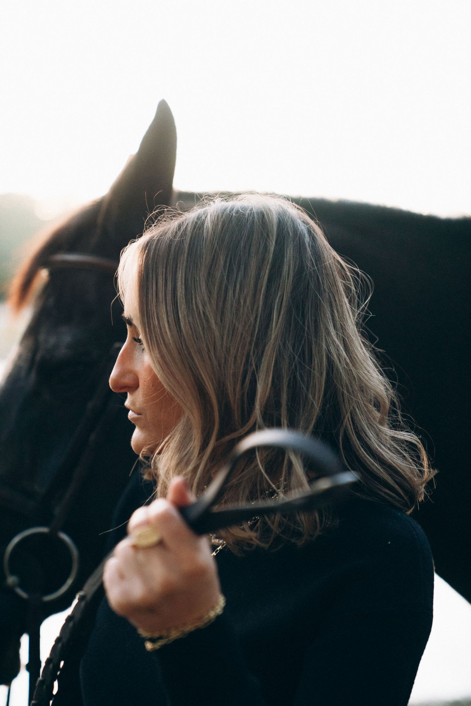 Side profile portrait of a person with wavy blonde hair standing next to a horse with bridle in soft lighting.