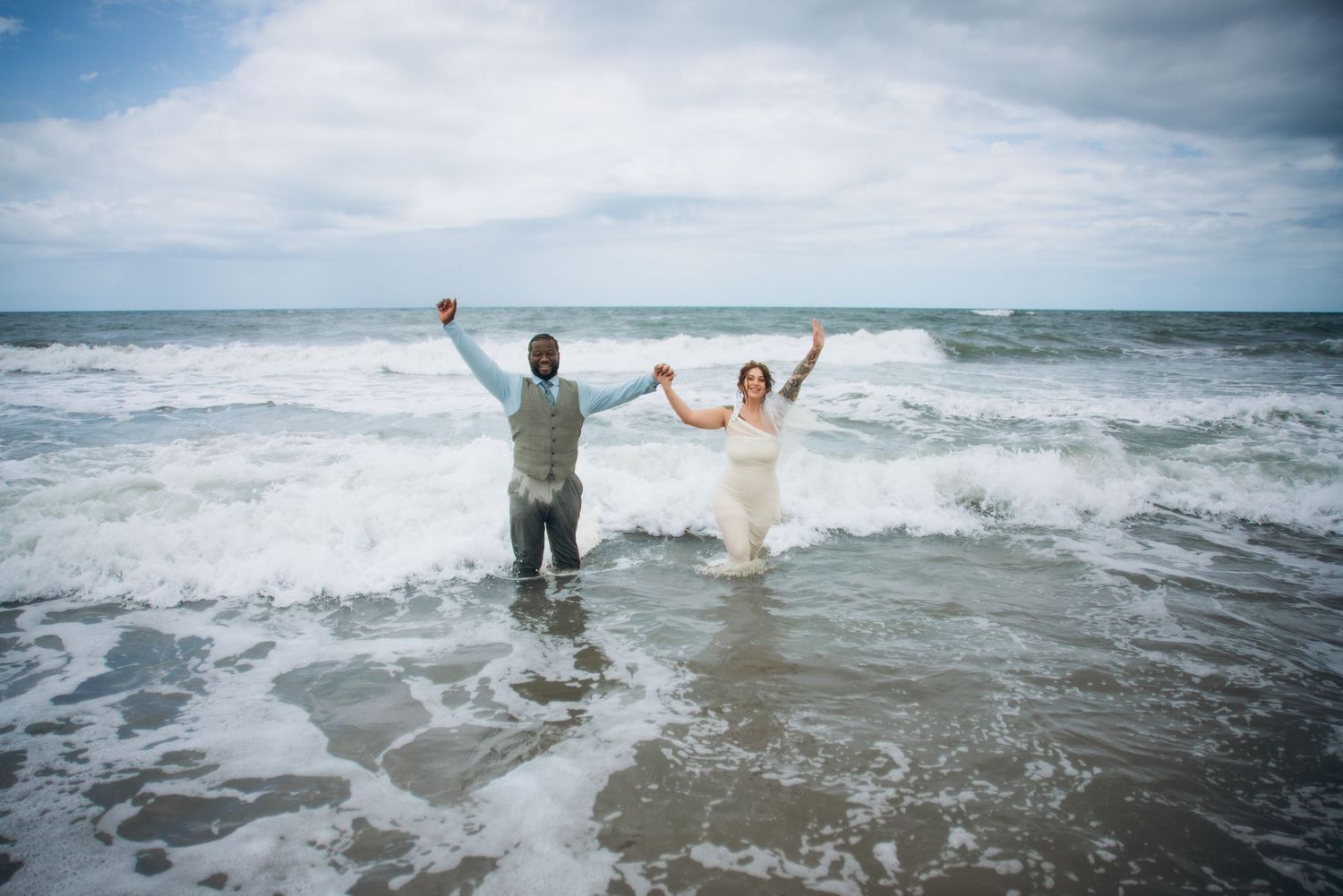 Joyful bride and groom standing in ocean waves with arms raised in celebration at their beach wedding.