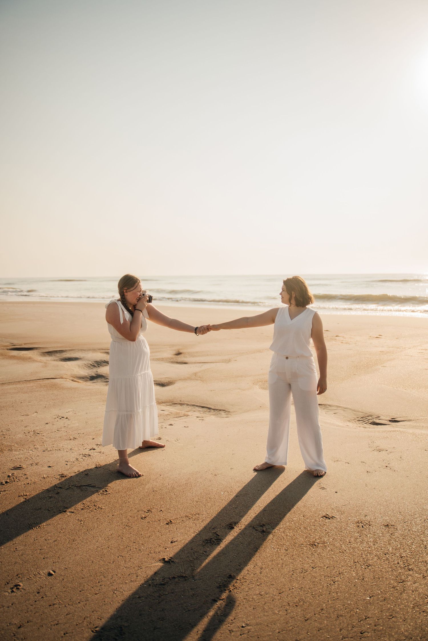 Two people in white outfits hold hands on a beach as long shadows stretch across golden sand.