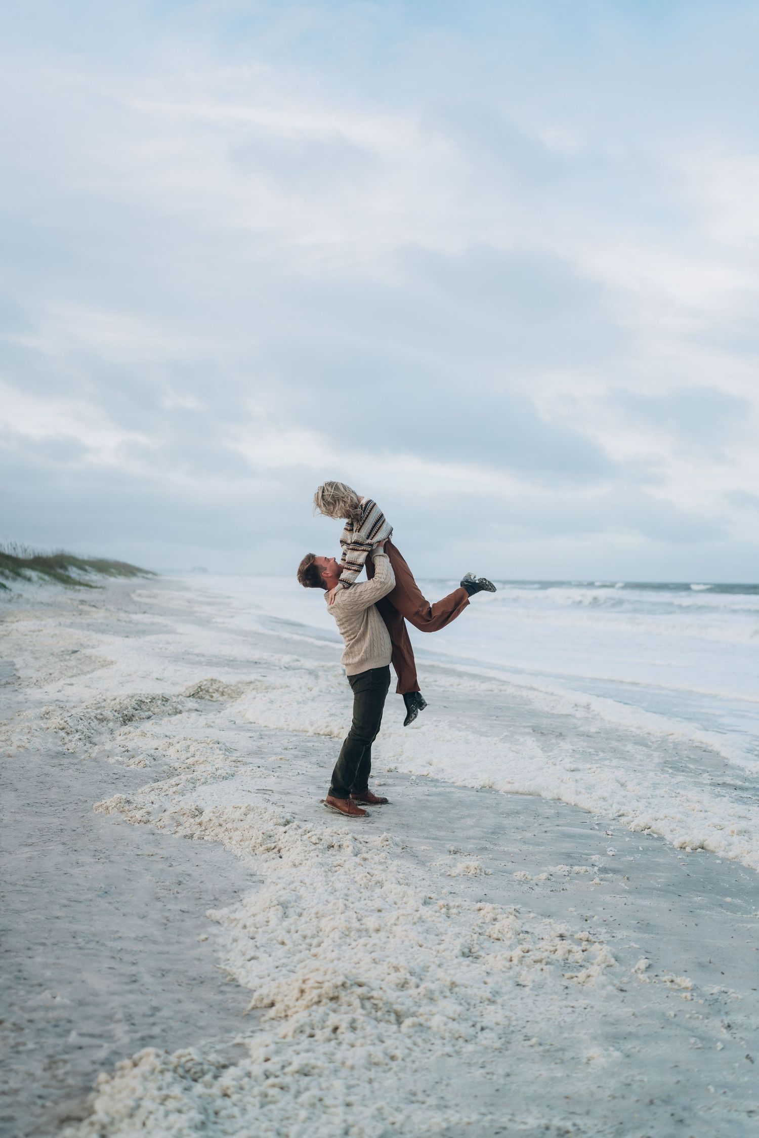 A romantic beach scene shows one person lifting another in a joyful embrace by the ocean.