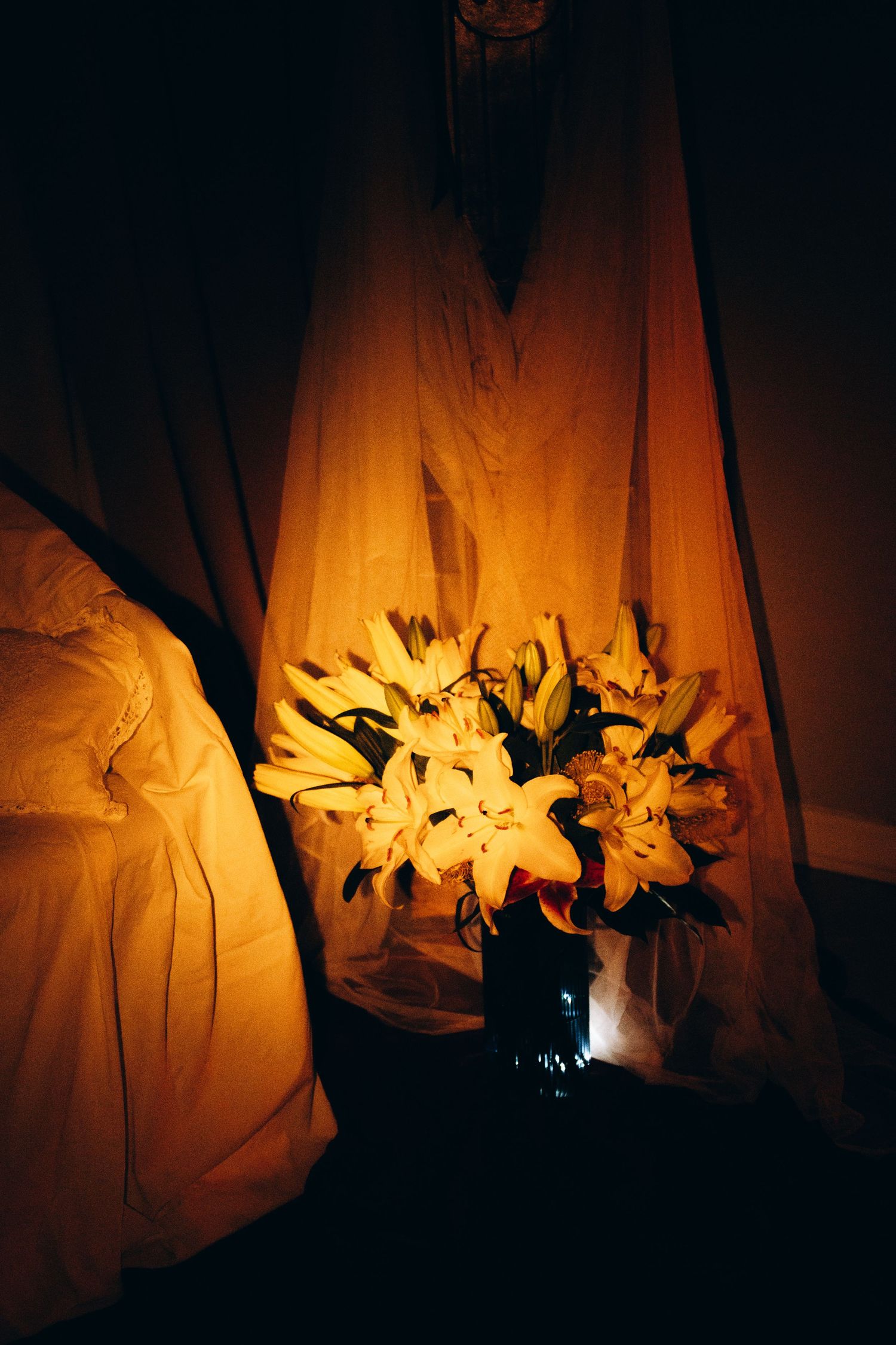 White lilies in a vase glowing warmly beside draped fabric curtains in a dimly lit romantic room.