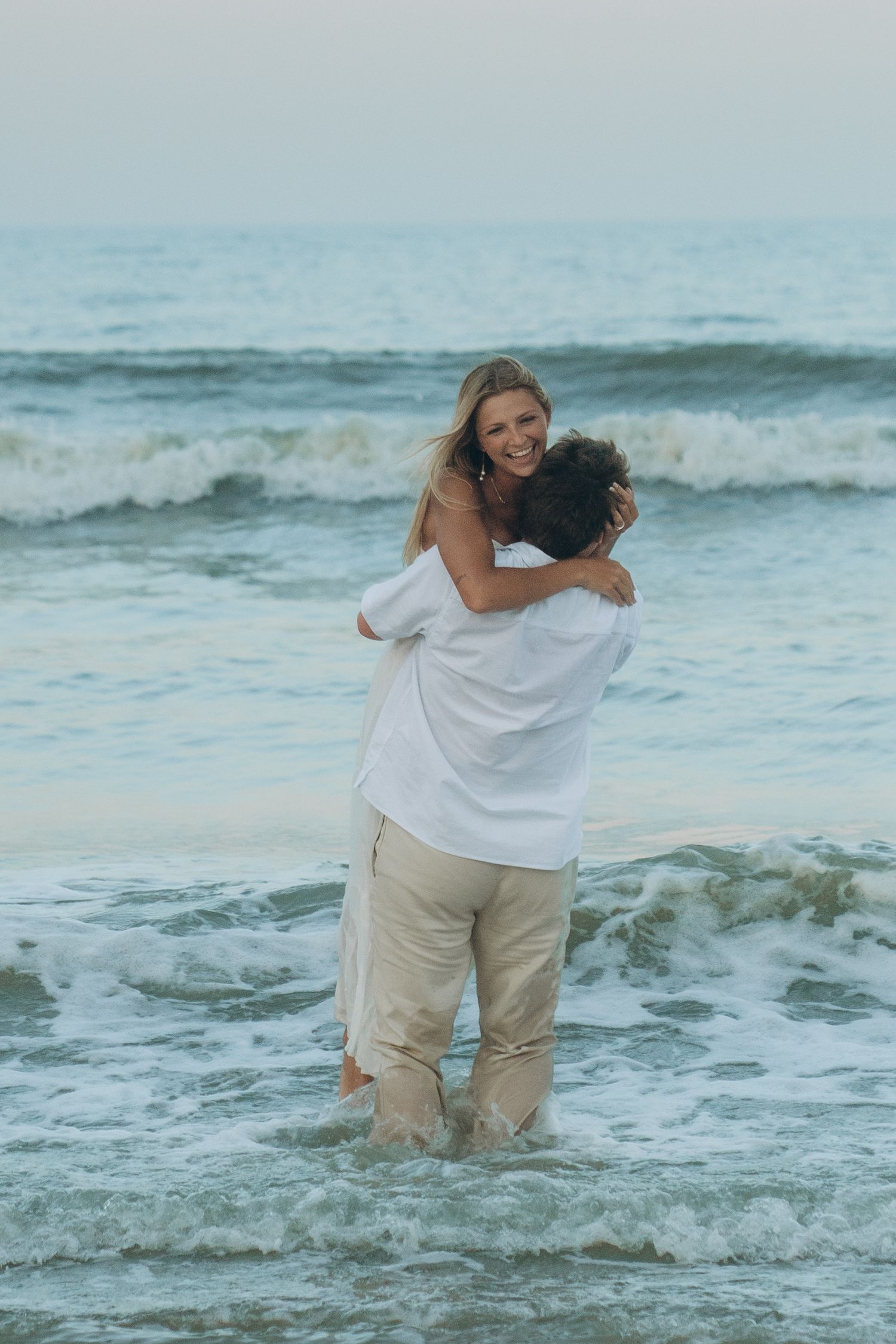 A couple in white and khaki clothing embraces while standing in the ocean waves.