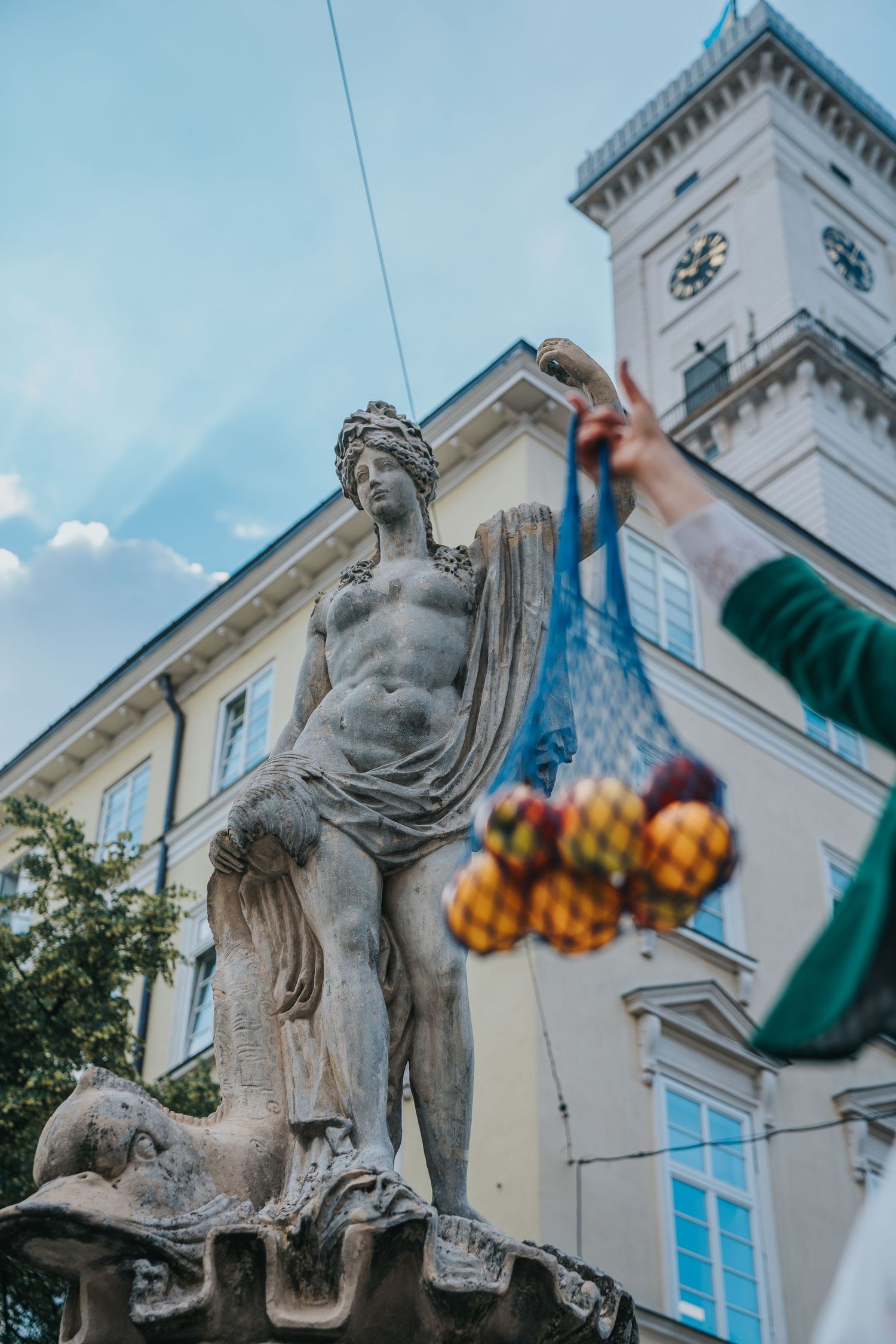 Classical stone statue in a European square with a clock tower and a hand holding a mesh bag of fruit.