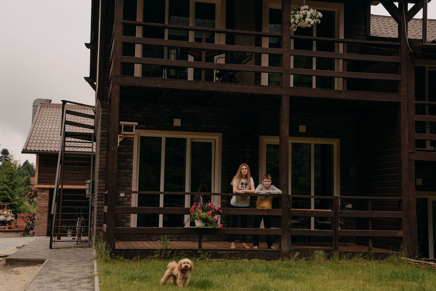 Two people relax on the porch of a dark wooden cabin while a golden dog plays on the green lawn below.
