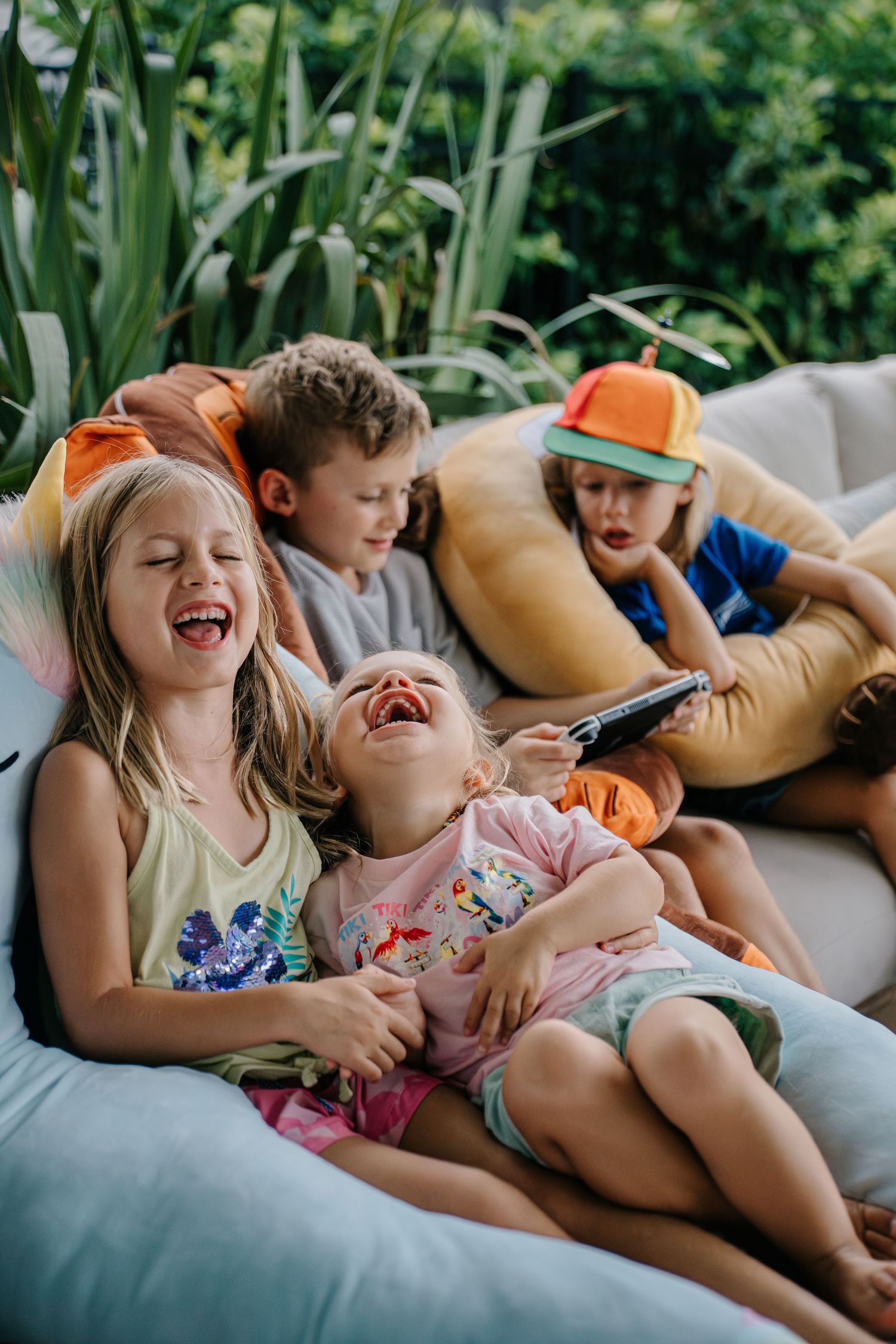 Four children laughing and relaxing together on outdoor cushions surrounded by lush green plants on a summer day.