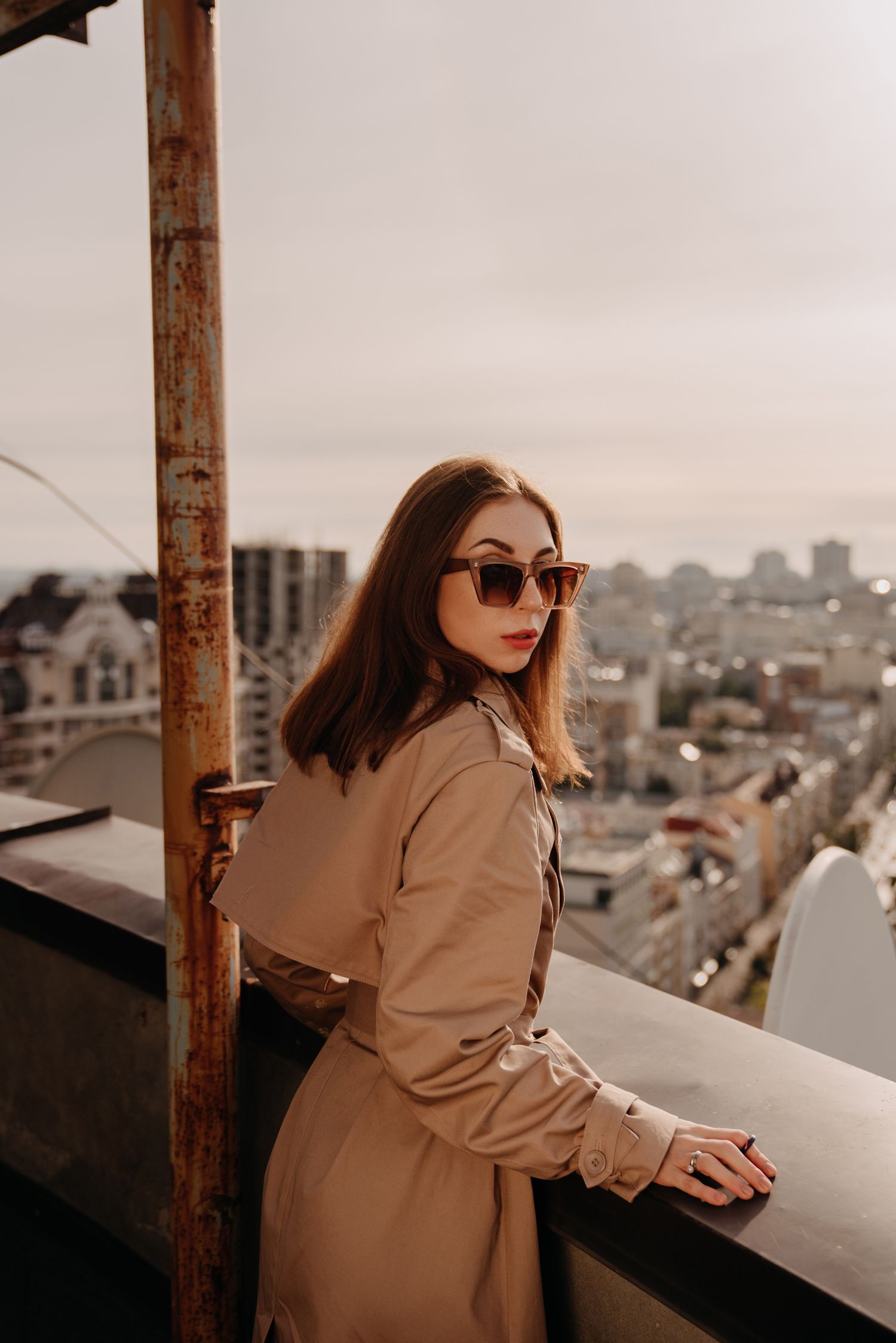 Young woman in beige trench coat and sunglasses poses on rooftop with city skyline backdrop.
