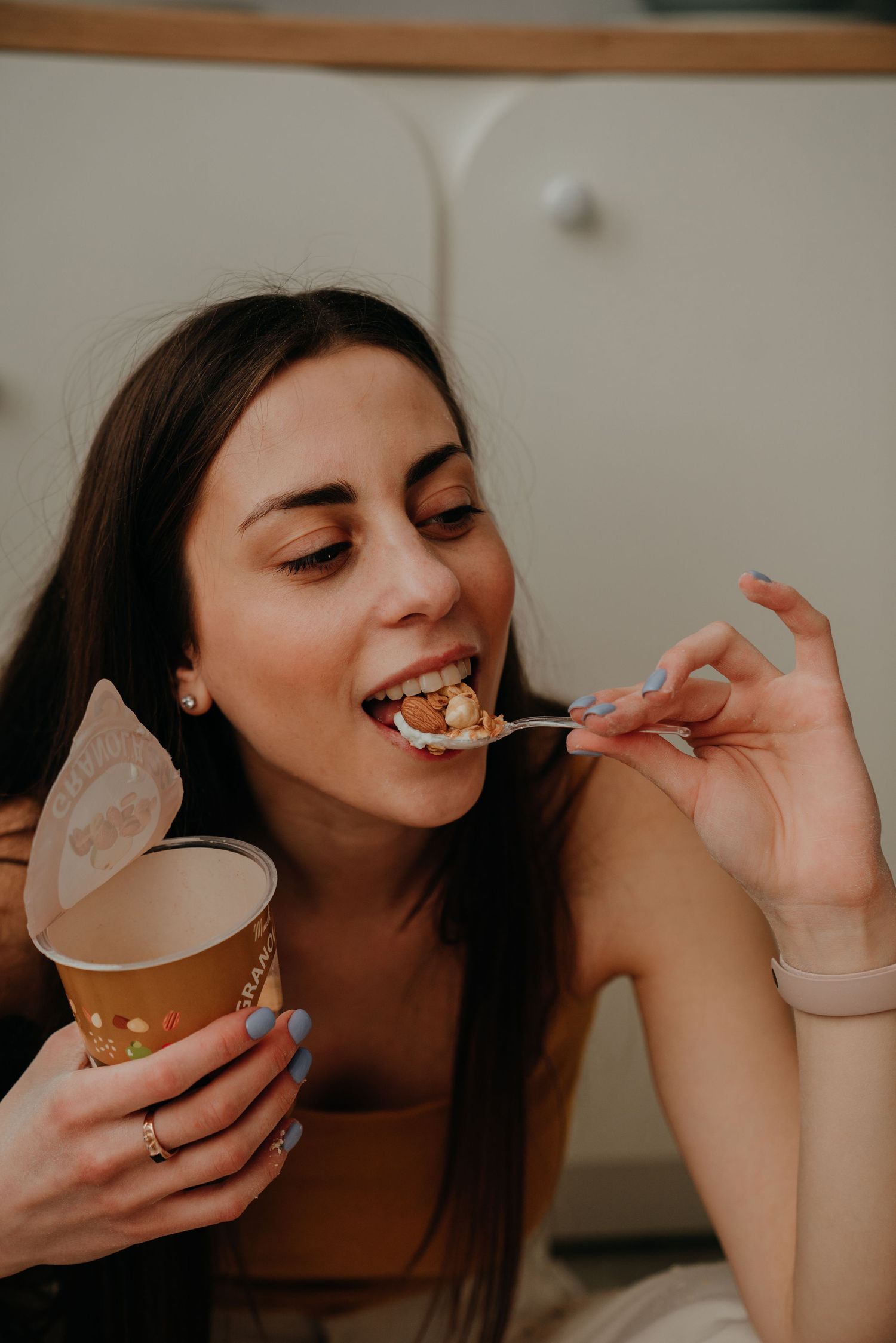 Young woman with blue nails enjoying a spoonful of granola from a paper cup, smiling with eyes closed.