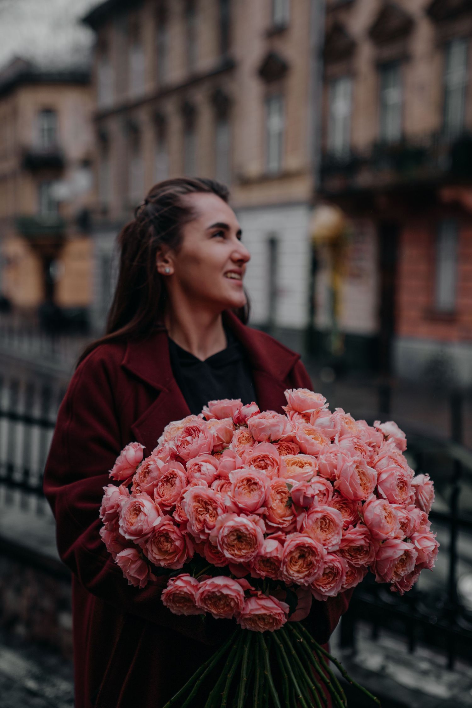 Smiling woman in burgundy coat holding large bouquet of pink roses on a city street.