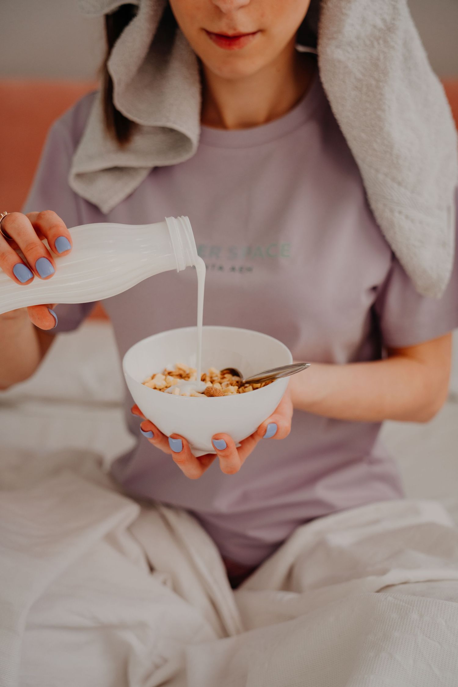 Woman pouring milk into a bowl of granola while sitting in bed with a towel on her head.