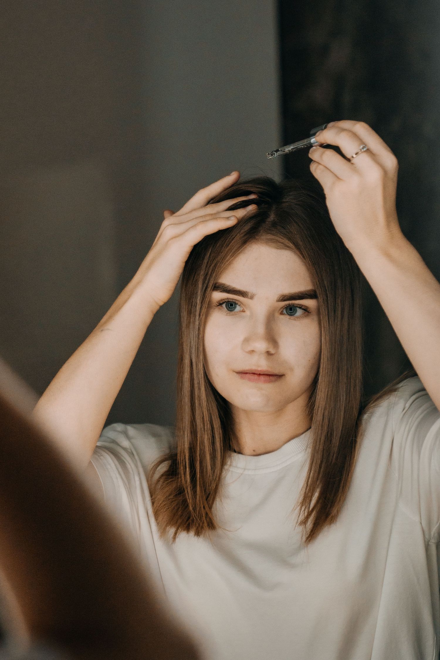 Young woman applying hair serum to roots while looking in mirror, wearing white t-shirt.